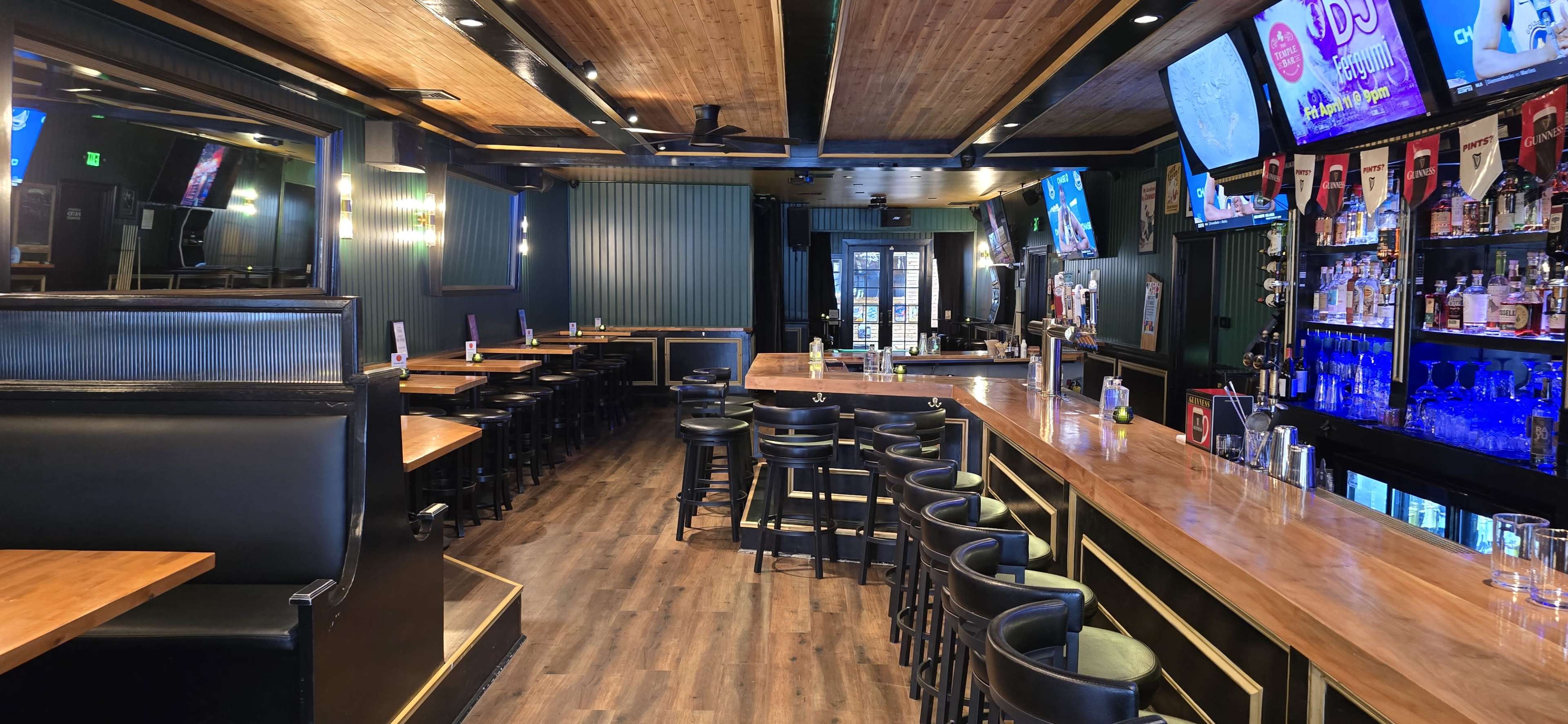 The image shows a modern bar interior with a wooden ceiling, empty seating areas, and a long counter lined with bar stools.