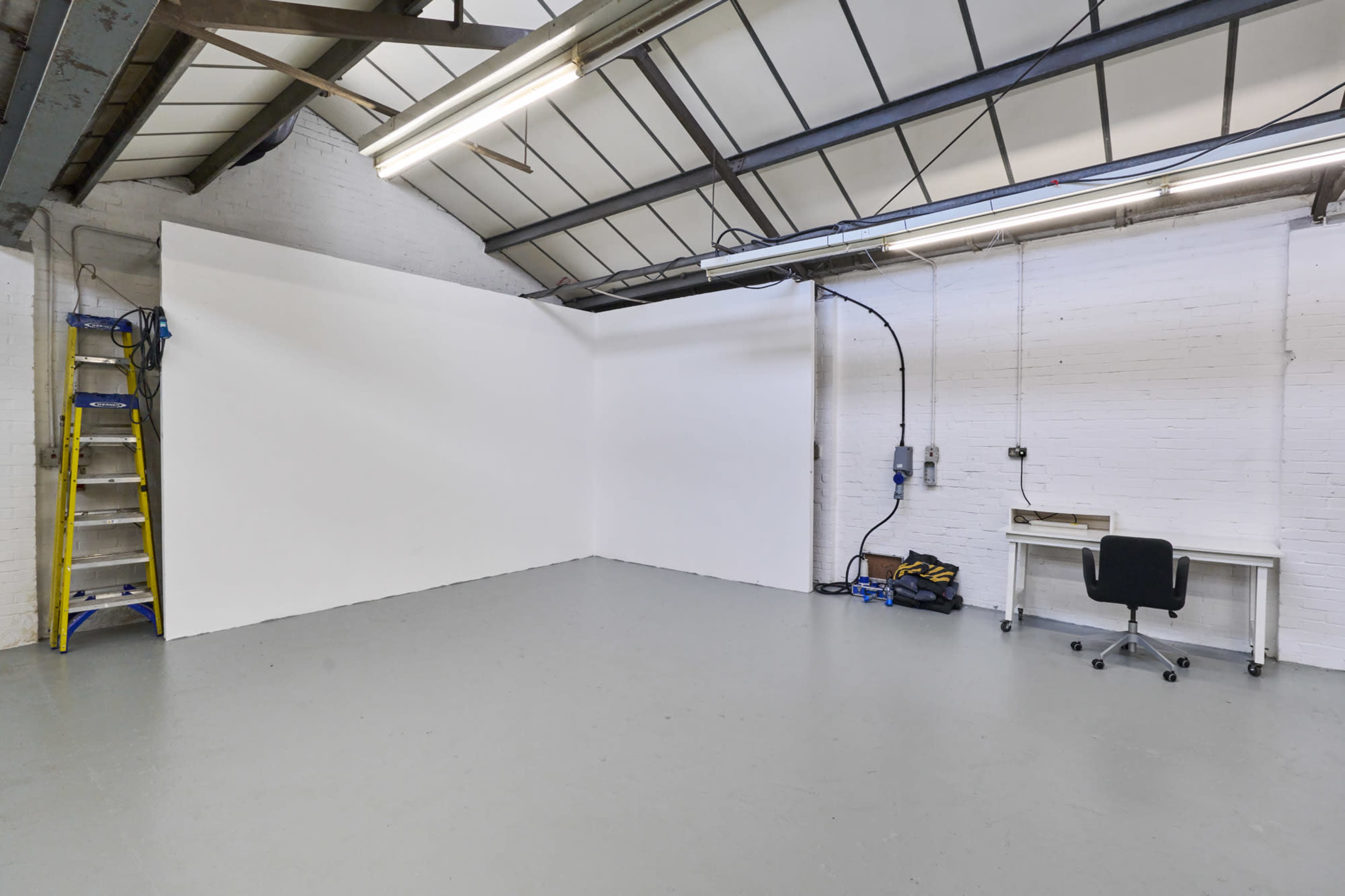 An empty industrial room with a white wall, a ladder, a desk, and a chair.