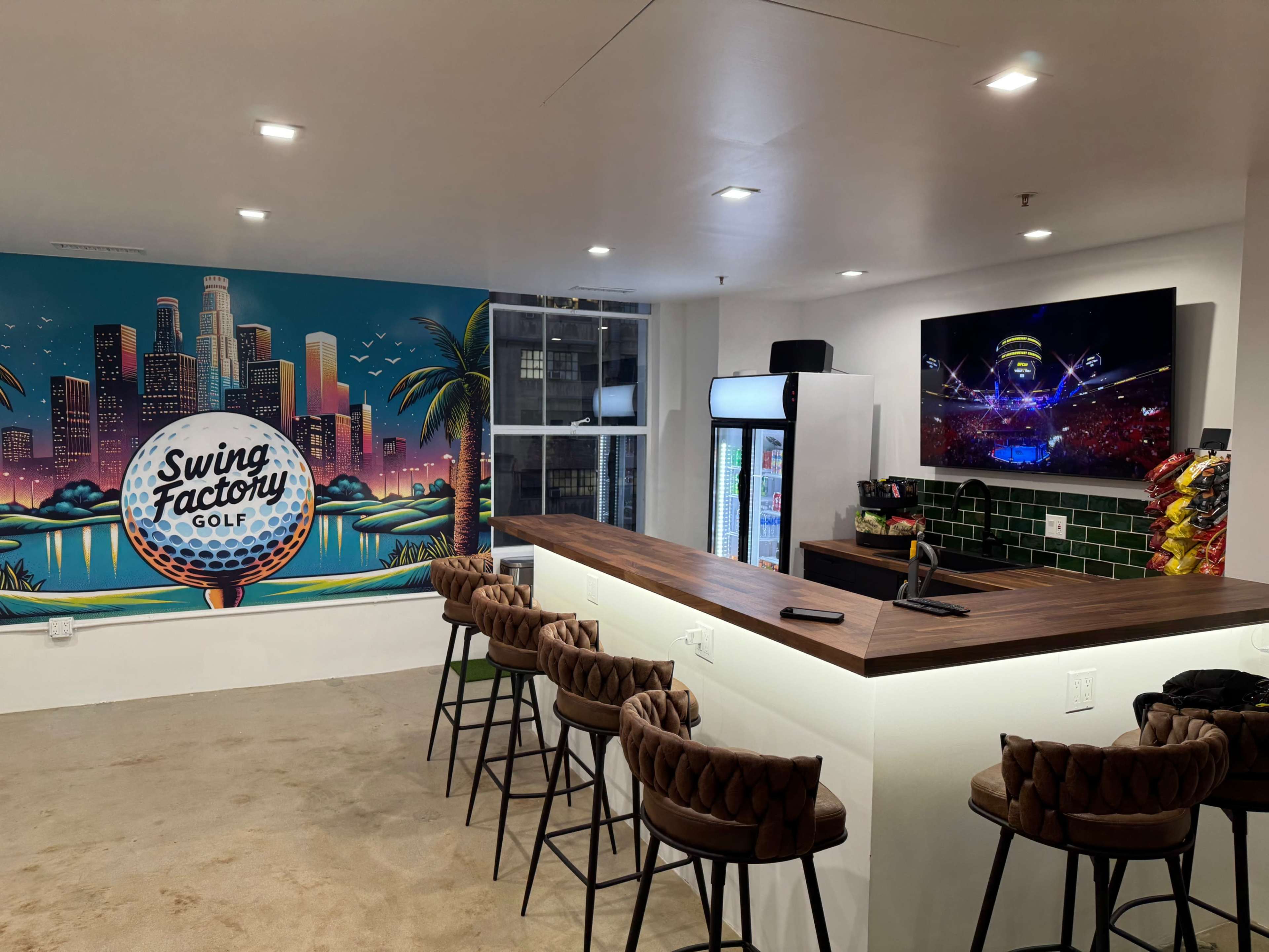 The image shows a modern indoor space featuring a bar with seating, a large television displaying a concert, and a colorful mural of a golf ball with city skyline and palm trees.