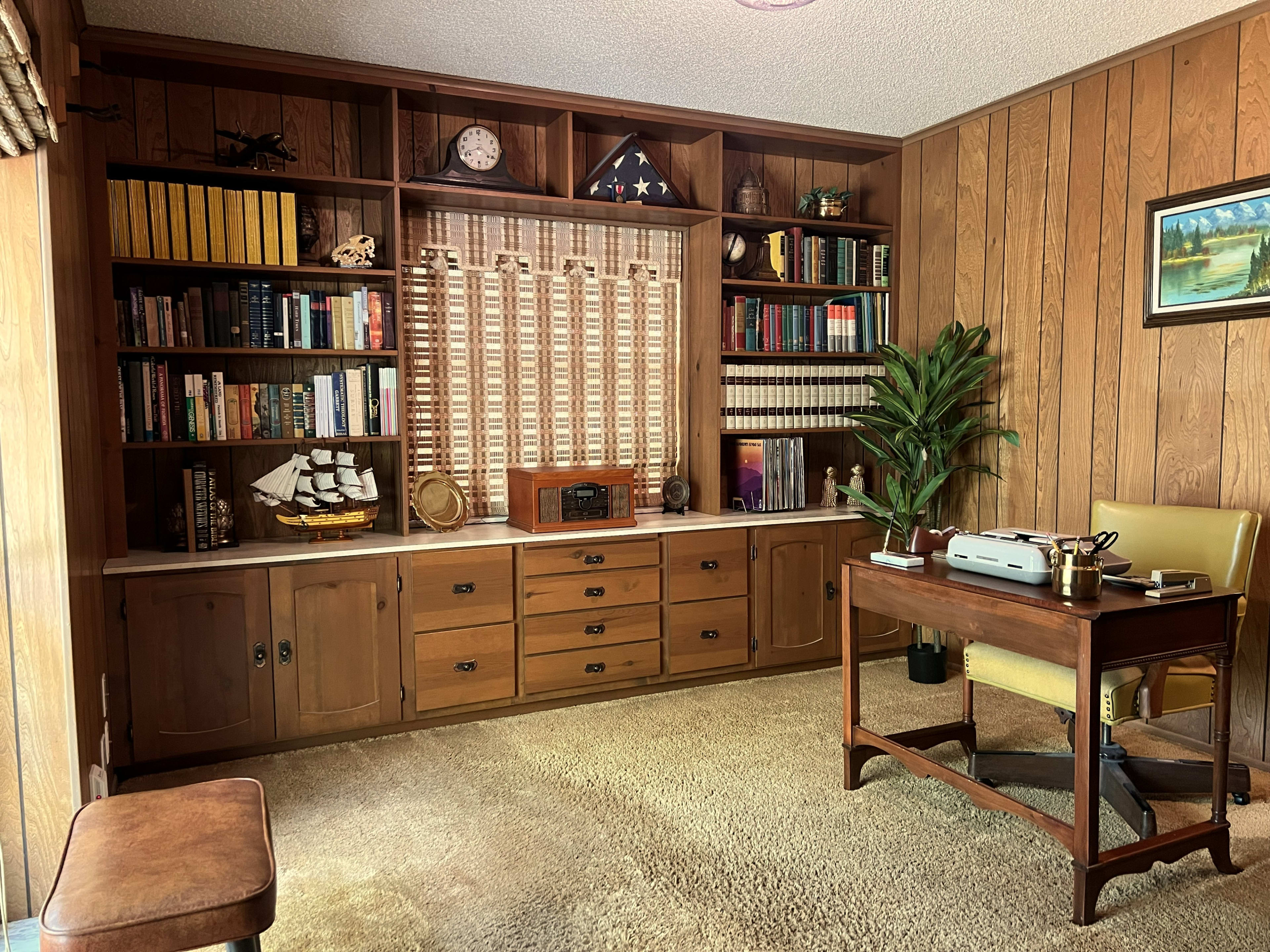 The image shows a wooden-paneled office with a bookshelf filled with books, a desk, a plant, and a vintage radio, all arranged in a cozy atmosphere.