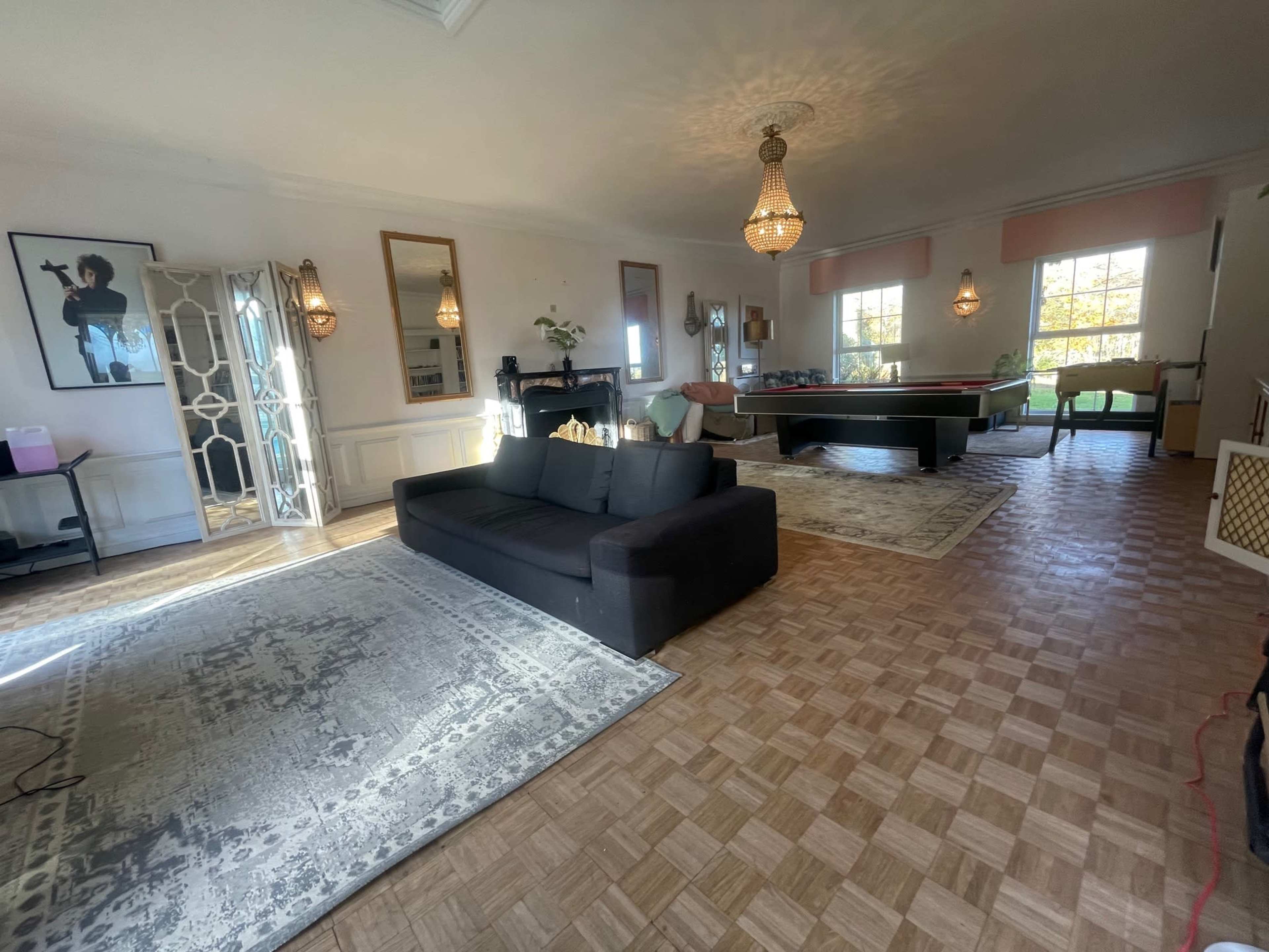 The image depicts a spacious living room featuring a dark sofa, a pool table, and multiple light fixtures, with large windows allowing natural light to enter.