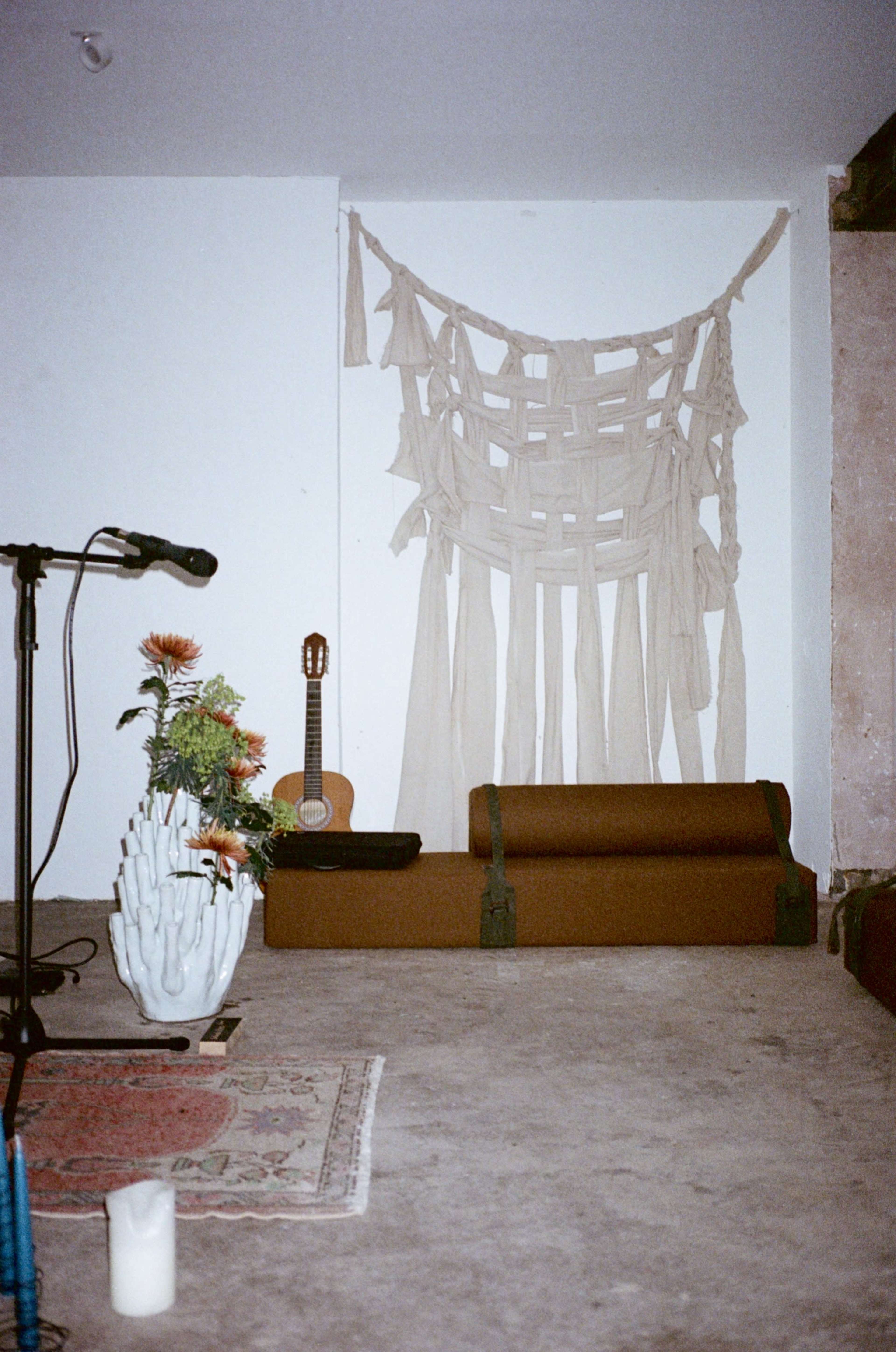 The scene features a minimalist room with a beige fabric wall hanging, a guitar on a stand, a decorative plant, and a candle on the floor.