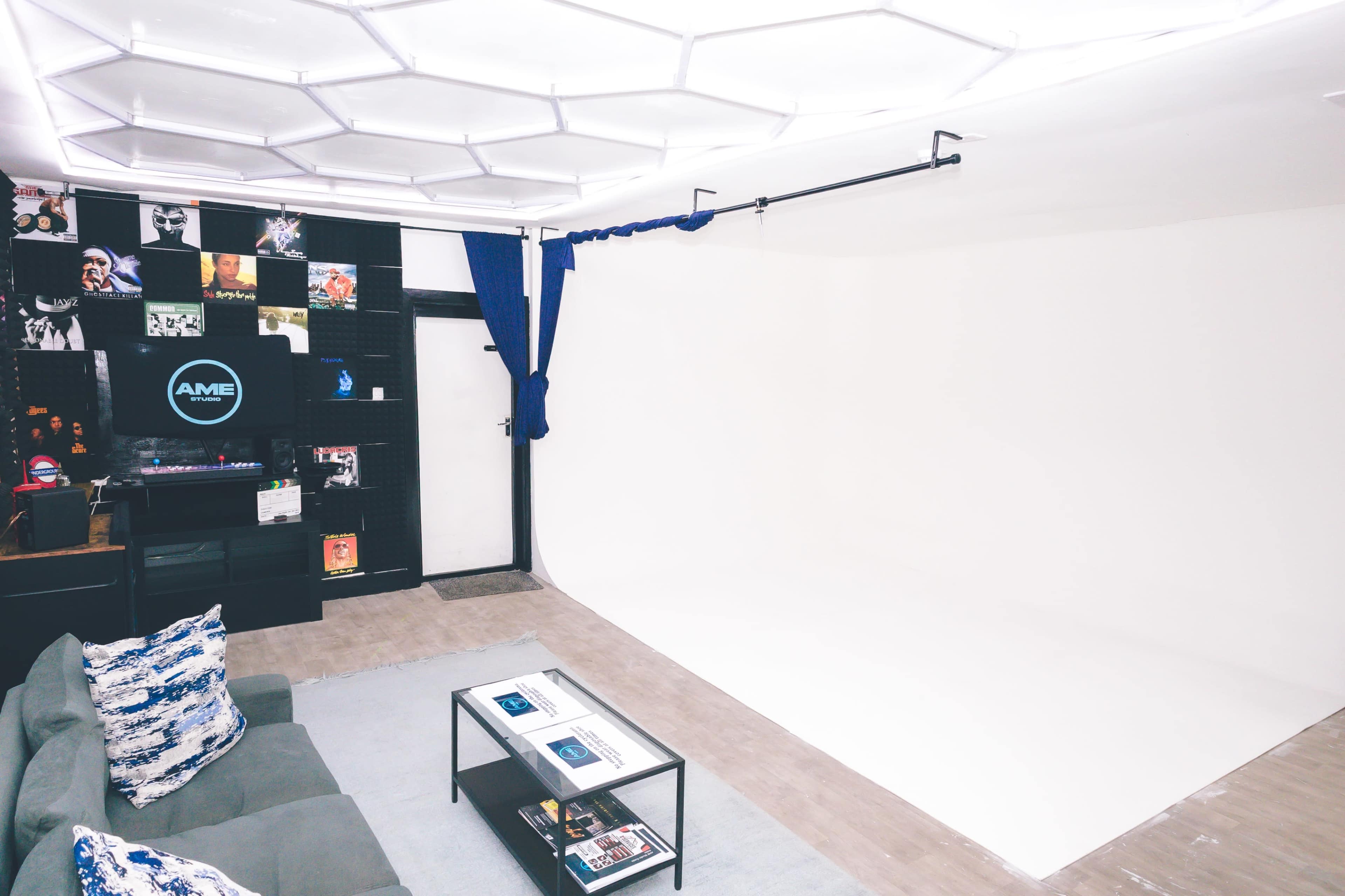 The image shows a minimalist studio space with a white background, a green sofa, a coffee table, and a wall decorated with black-and-white photographs and various artworks.