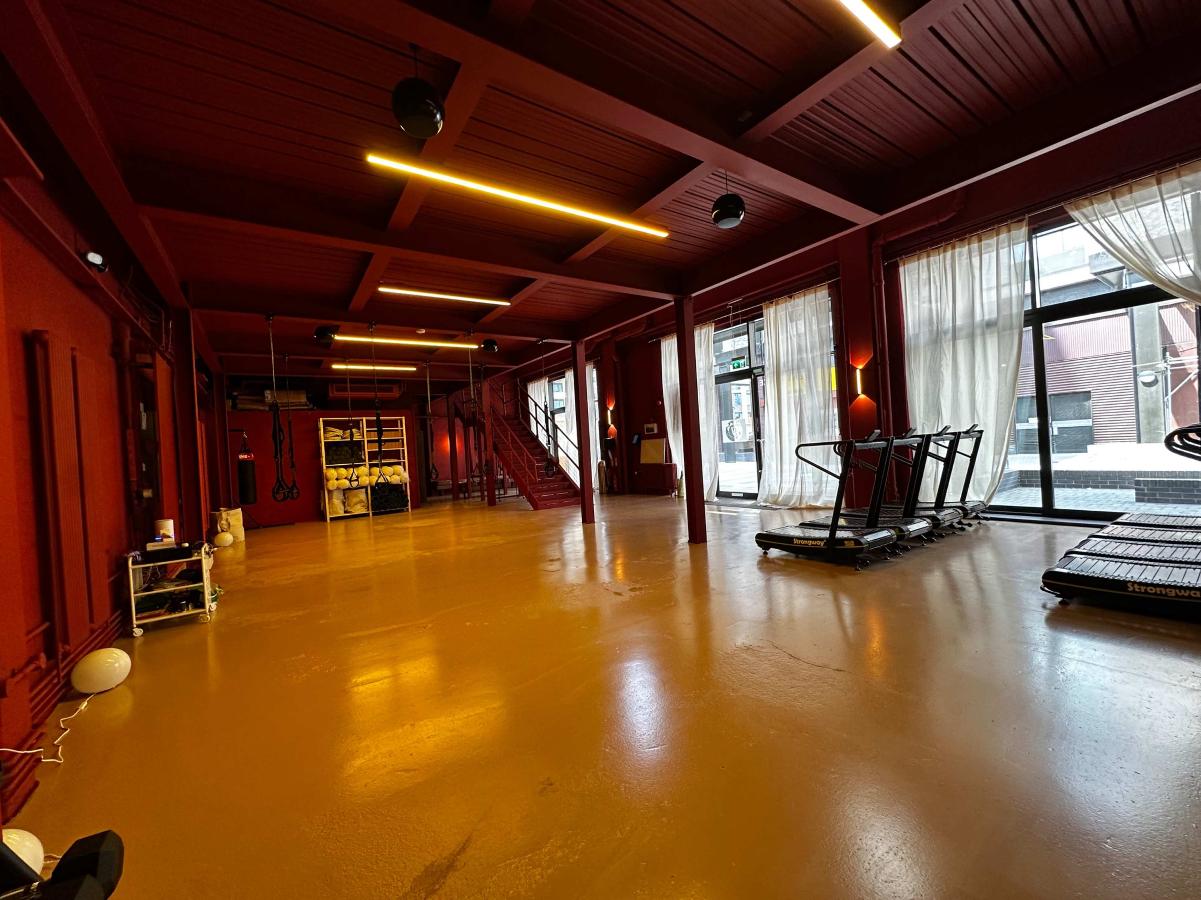 The image shows a spacious gym with red walls, exercise equipment, and large windows allowing natural light to enter.
