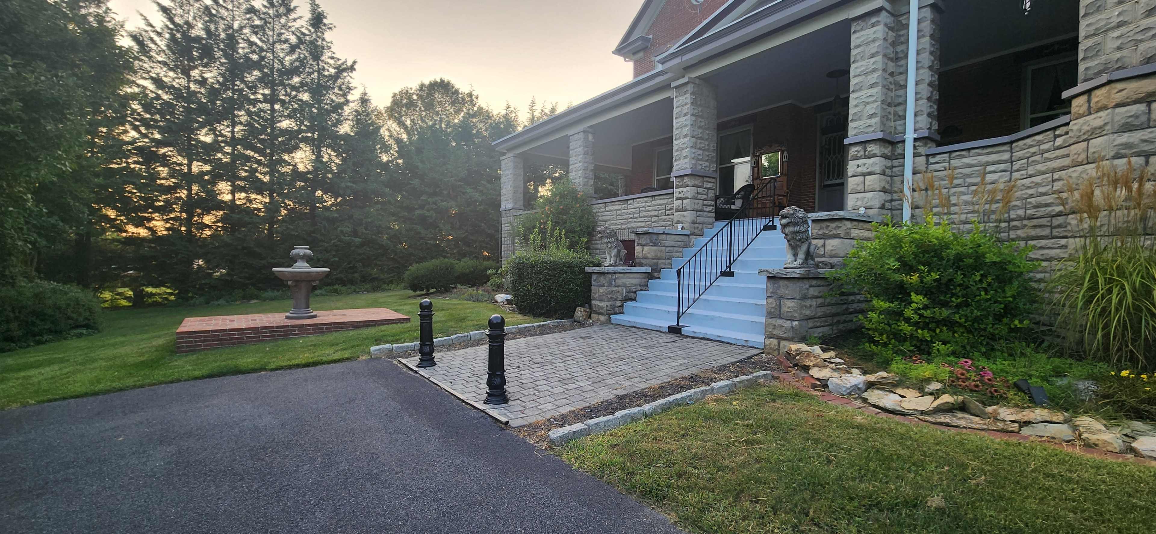 A stone porch with stairs leading to a brick pathway, flanked by bushes and decorative plants, in front of a house surrounded by trees.