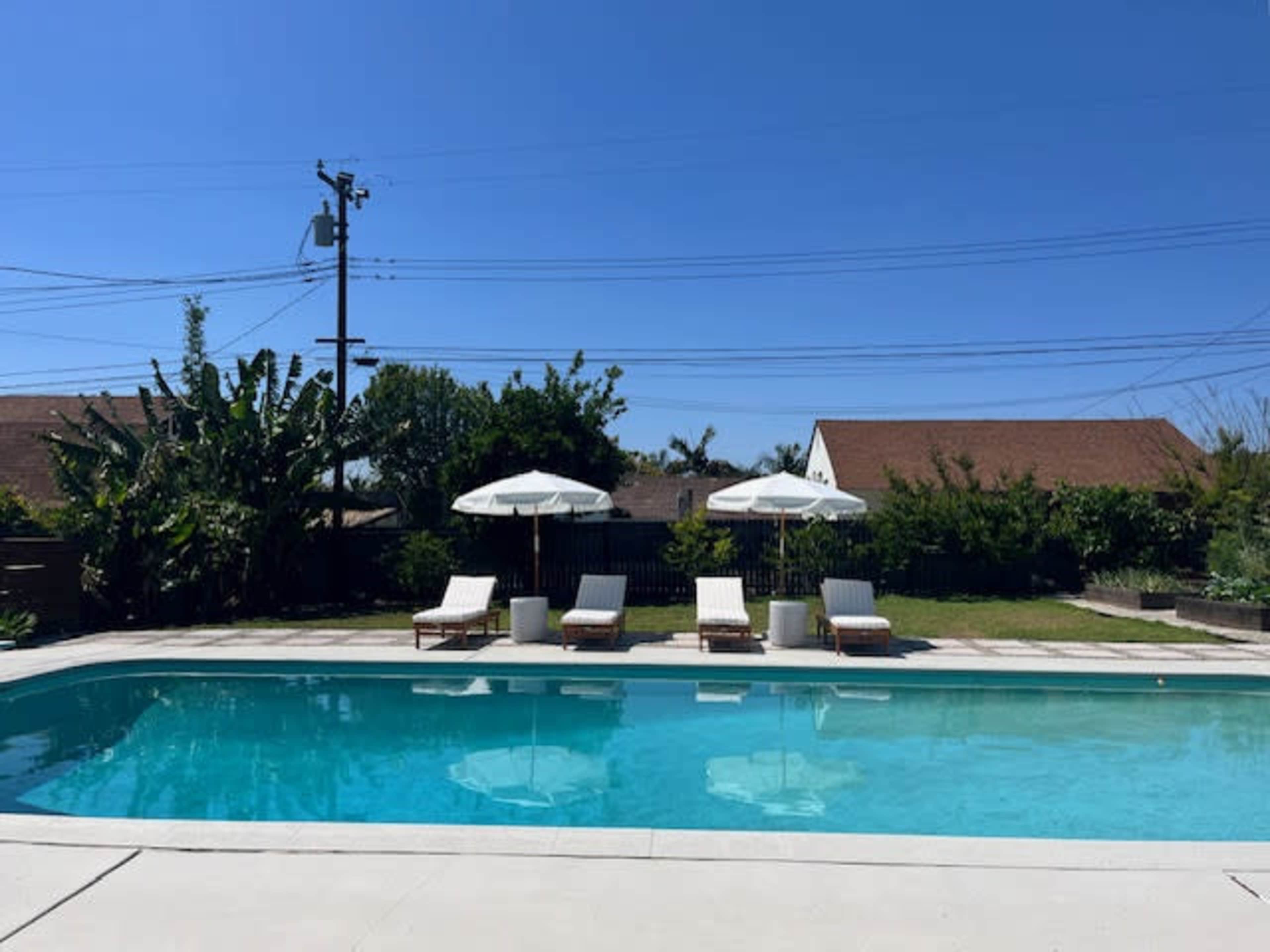 The image shows a clear swimming pool with three lounge chairs and two umbrellas beside it, surrounded by greenery under a bright blue sky.