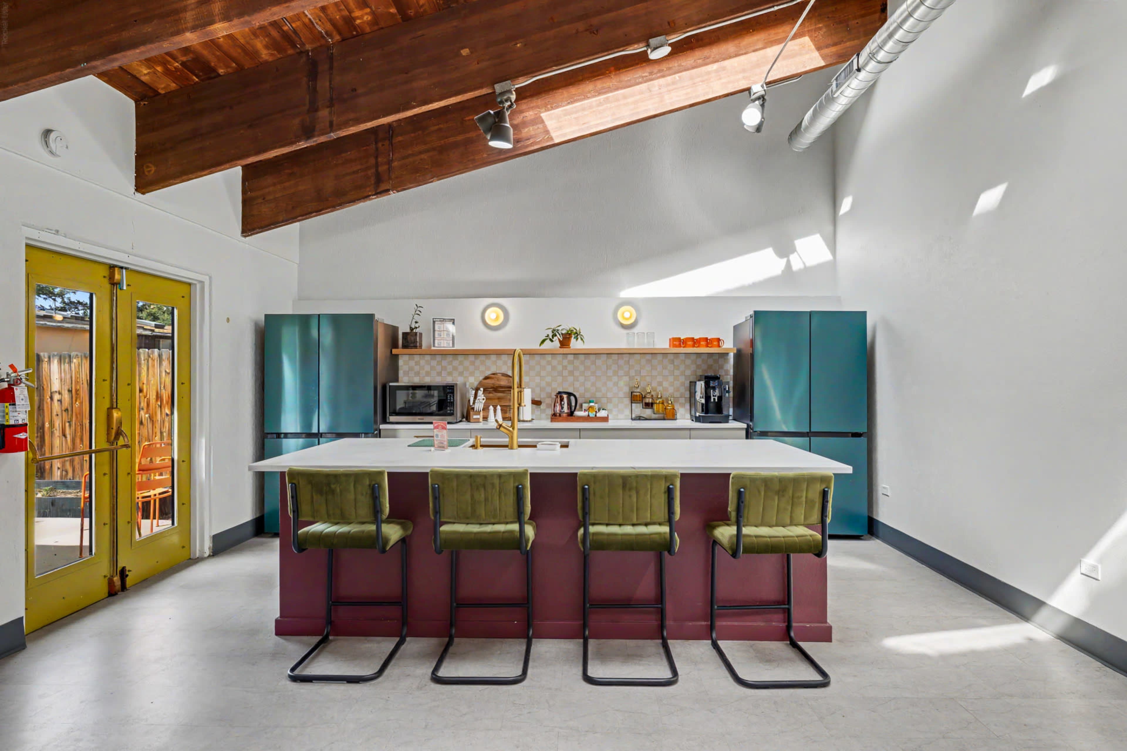 The image shows a modern kitchen with a large island bar, green stools, and blue appliances under a high wooden ceiling.