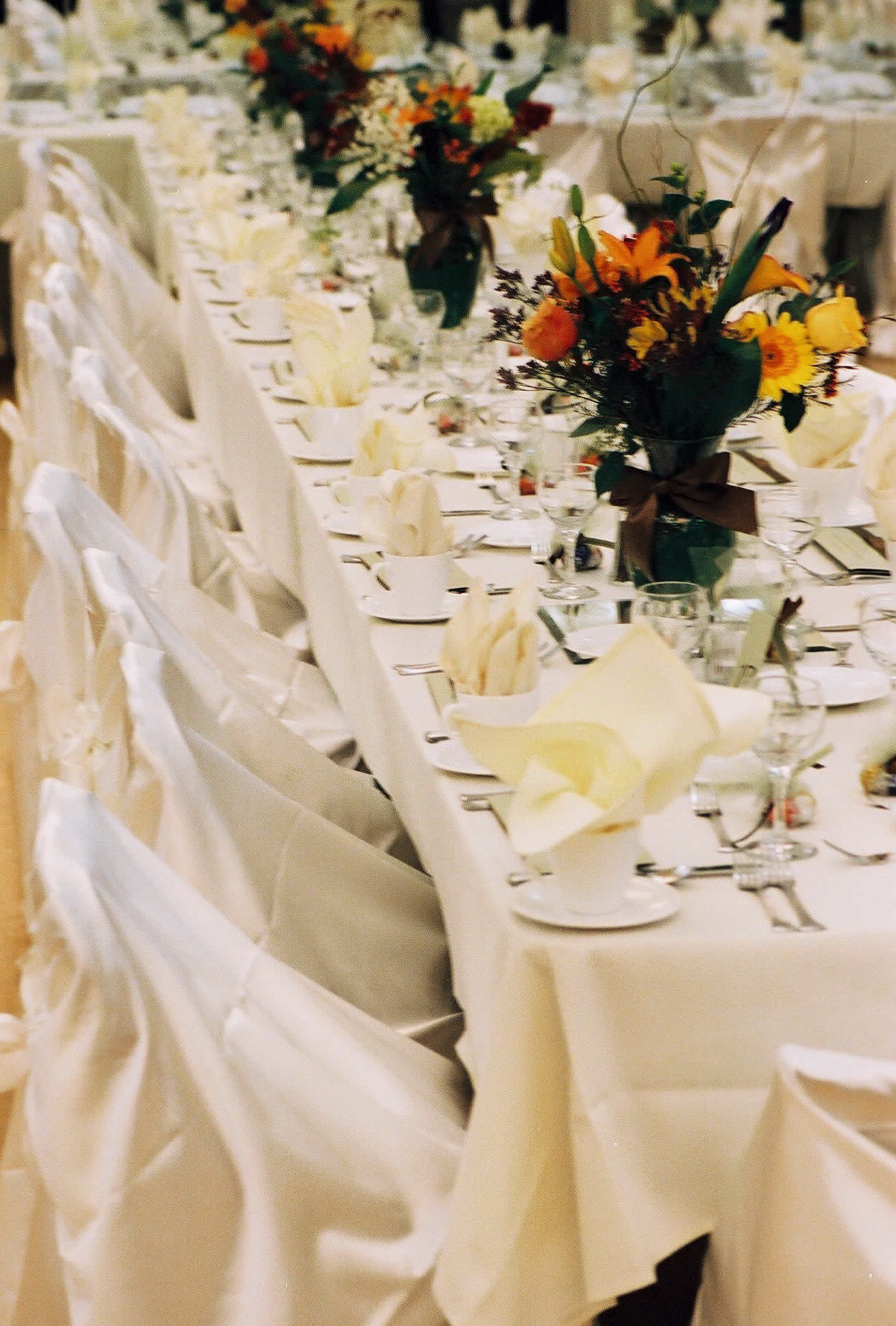 A long banquet table is elegantly set with white tablecloths, folded napkins, glassware, and floral centerpieces.