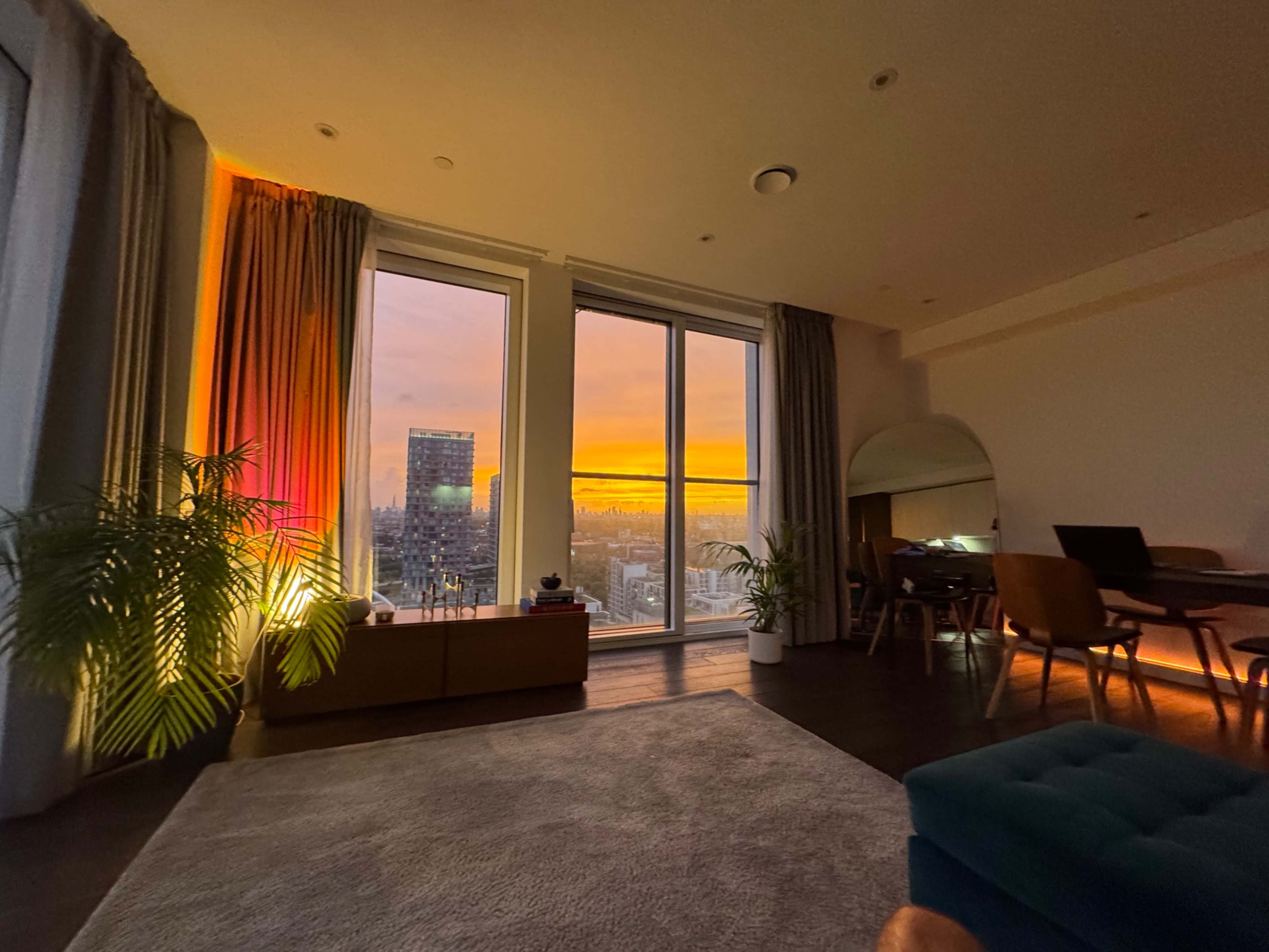 A modern living room with large windows showcases a vibrant sunset, illuminating the space with warm colors.