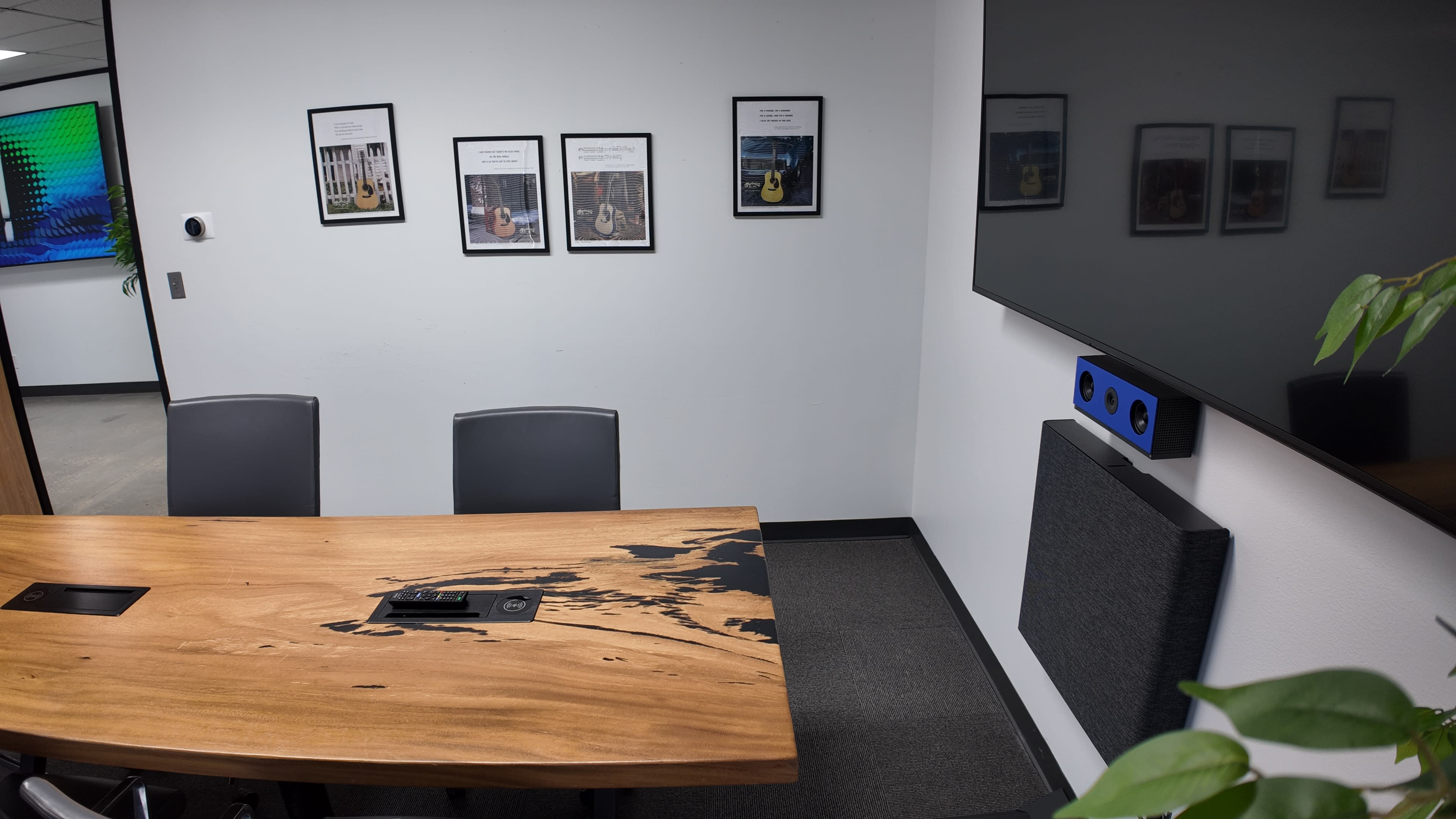 The image shows a conference room with a wooden table, black chairs, framed pictures on the wall, and a large screen mounted on the opposite wall.