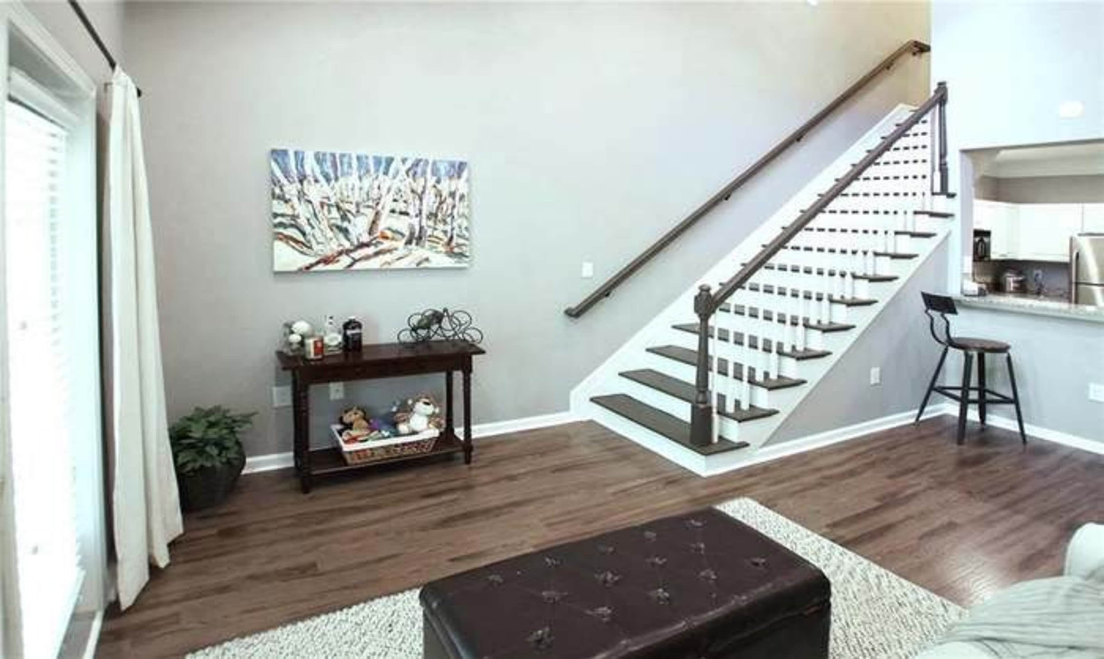 A well-lit living area with a staircase leading to an upper level, a small table against the wall, and a textured rug on the hardwood floor.