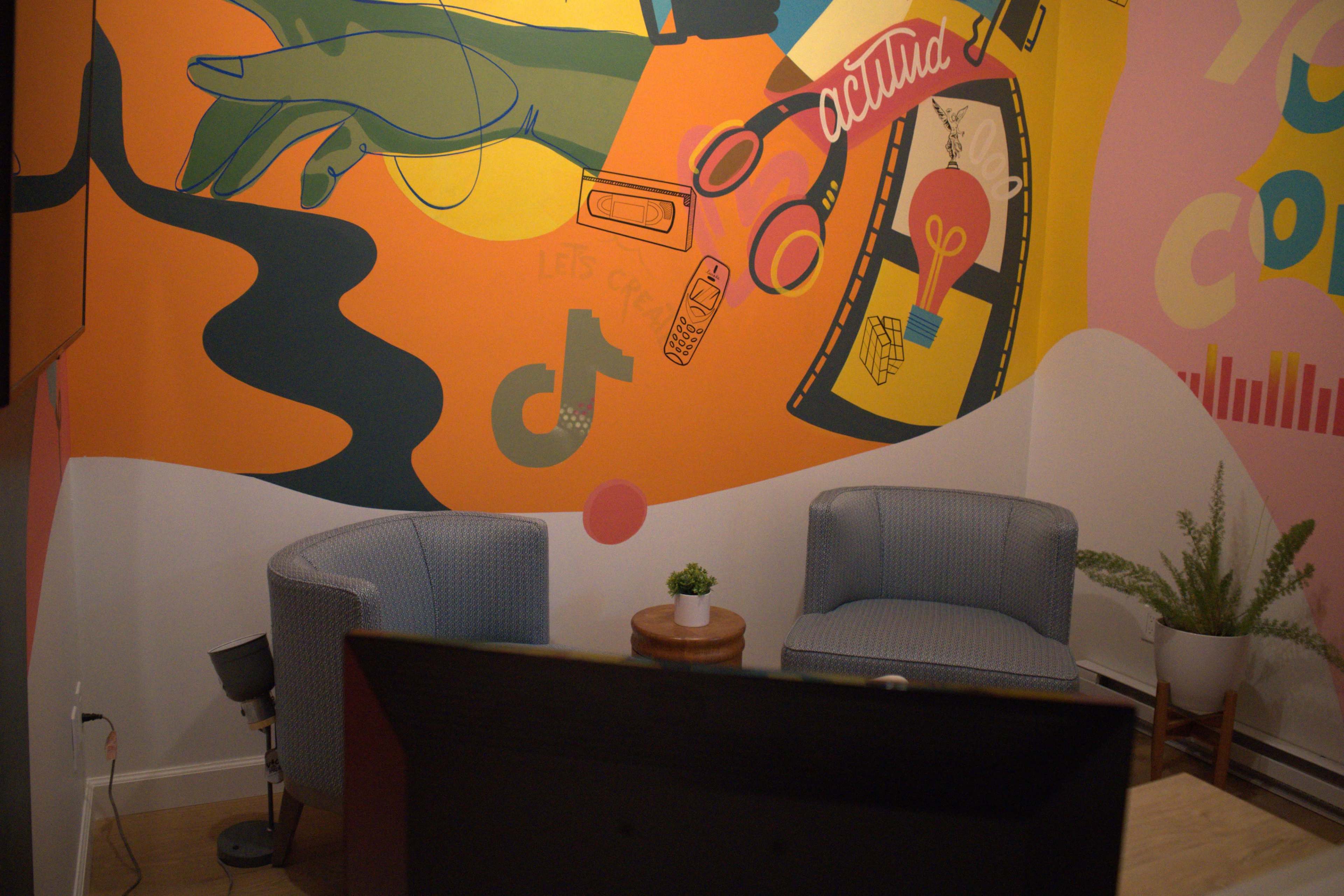 The image shows a colorful mural on a wall behind two blue chairs arranged around a small round table in a cozy room.