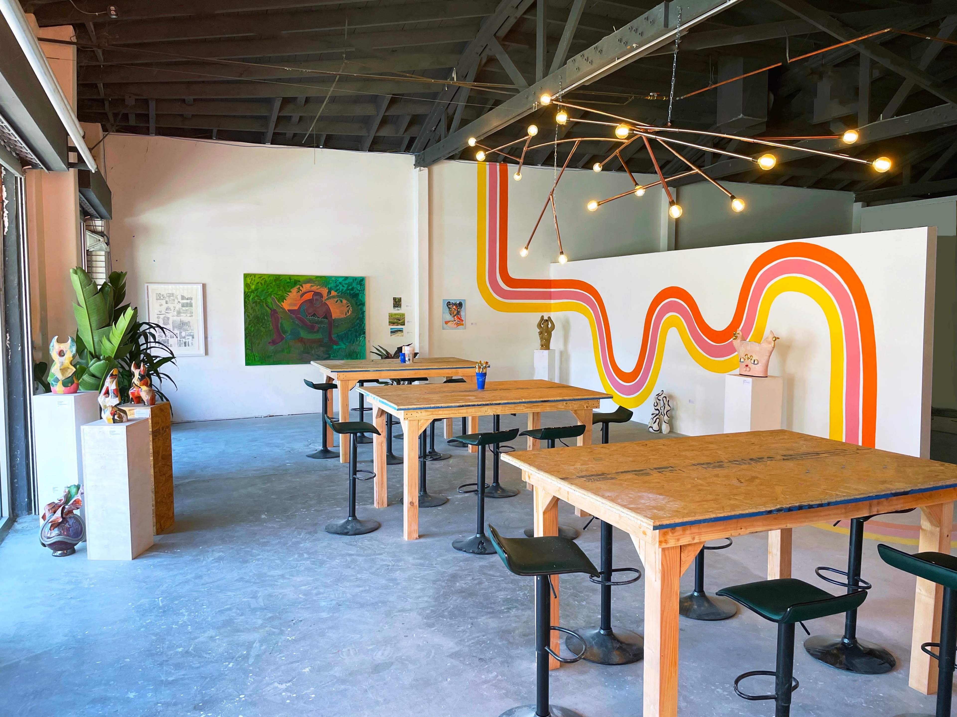 A spacious art studio with wooden tables and colorful wall art featuring bold orange and yellow lines.