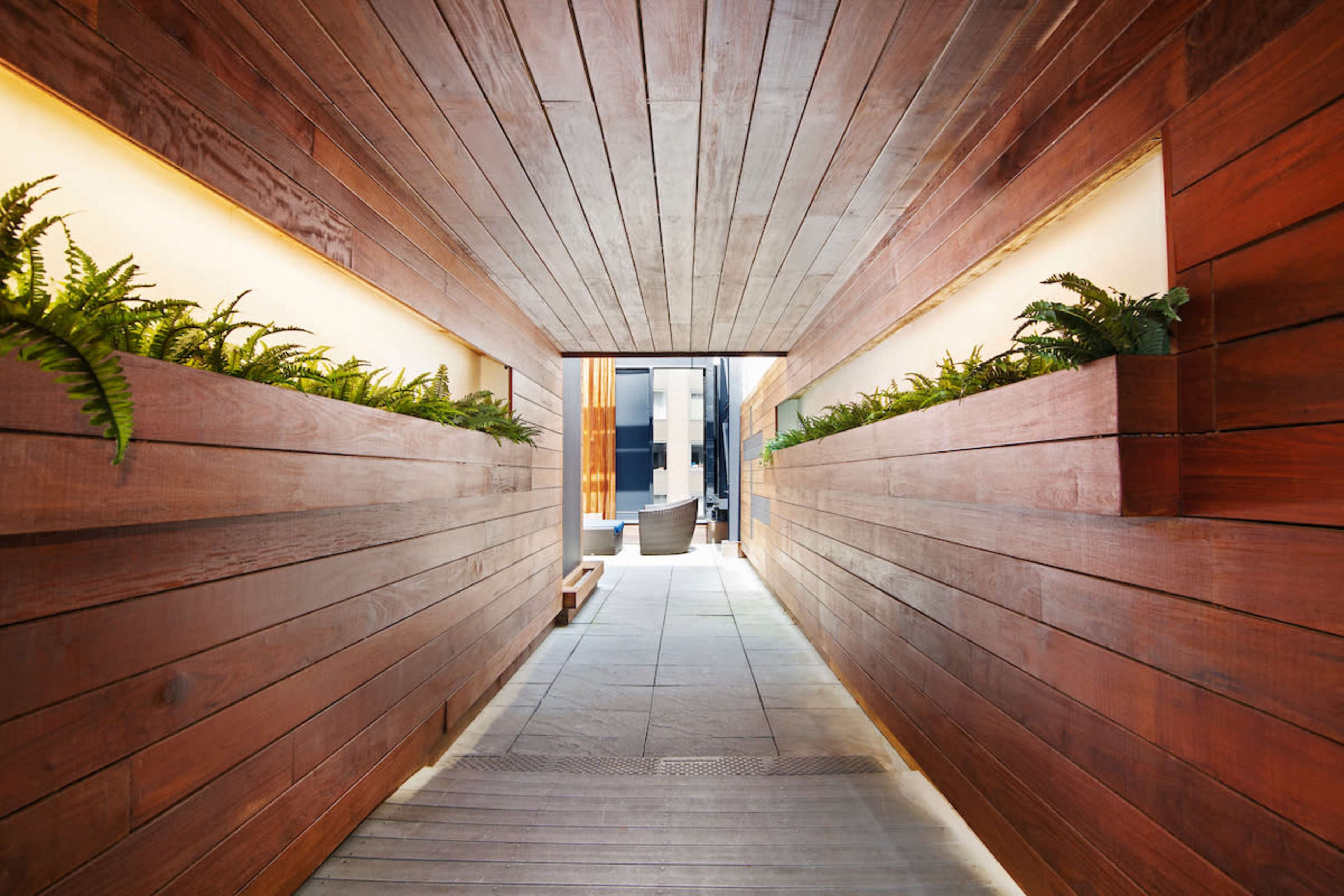 The image depicts a long, narrow hallway lined with wooden walls and illuminated by soft lighting, featuring ferns planted in raised beds along each side.