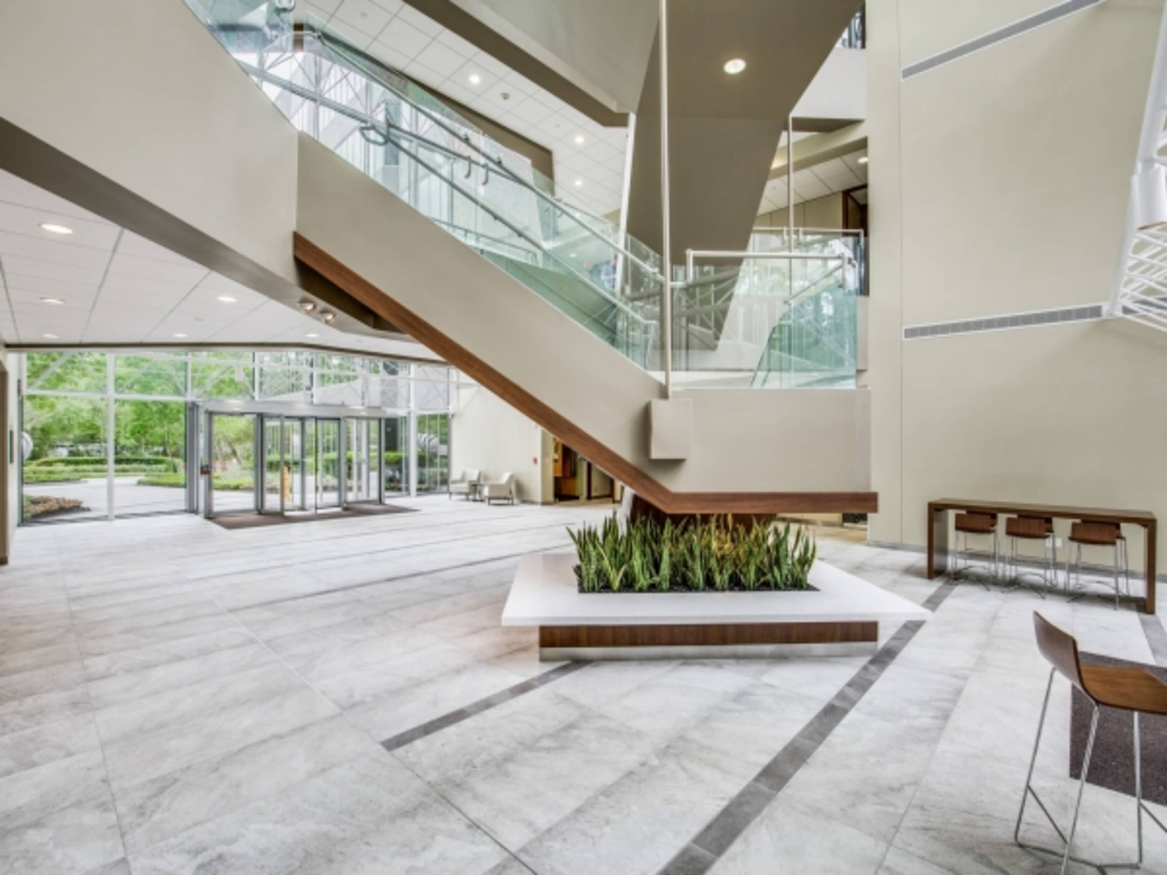 A modern lobby with a staircase, large windows, and a planter in the center of a tiled floor.
