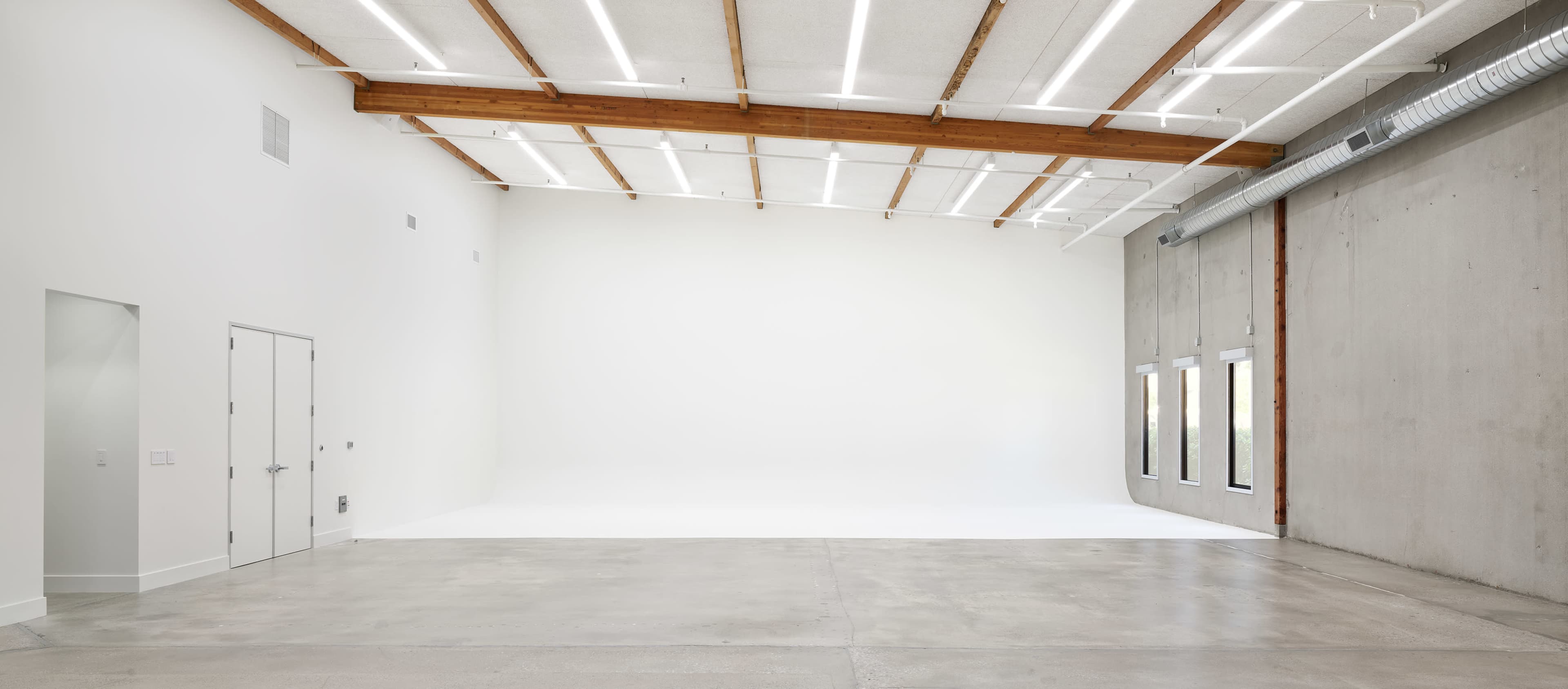 The image shows a spacious, minimalist studio with a concrete wall, wooden beams, and bright lighting.