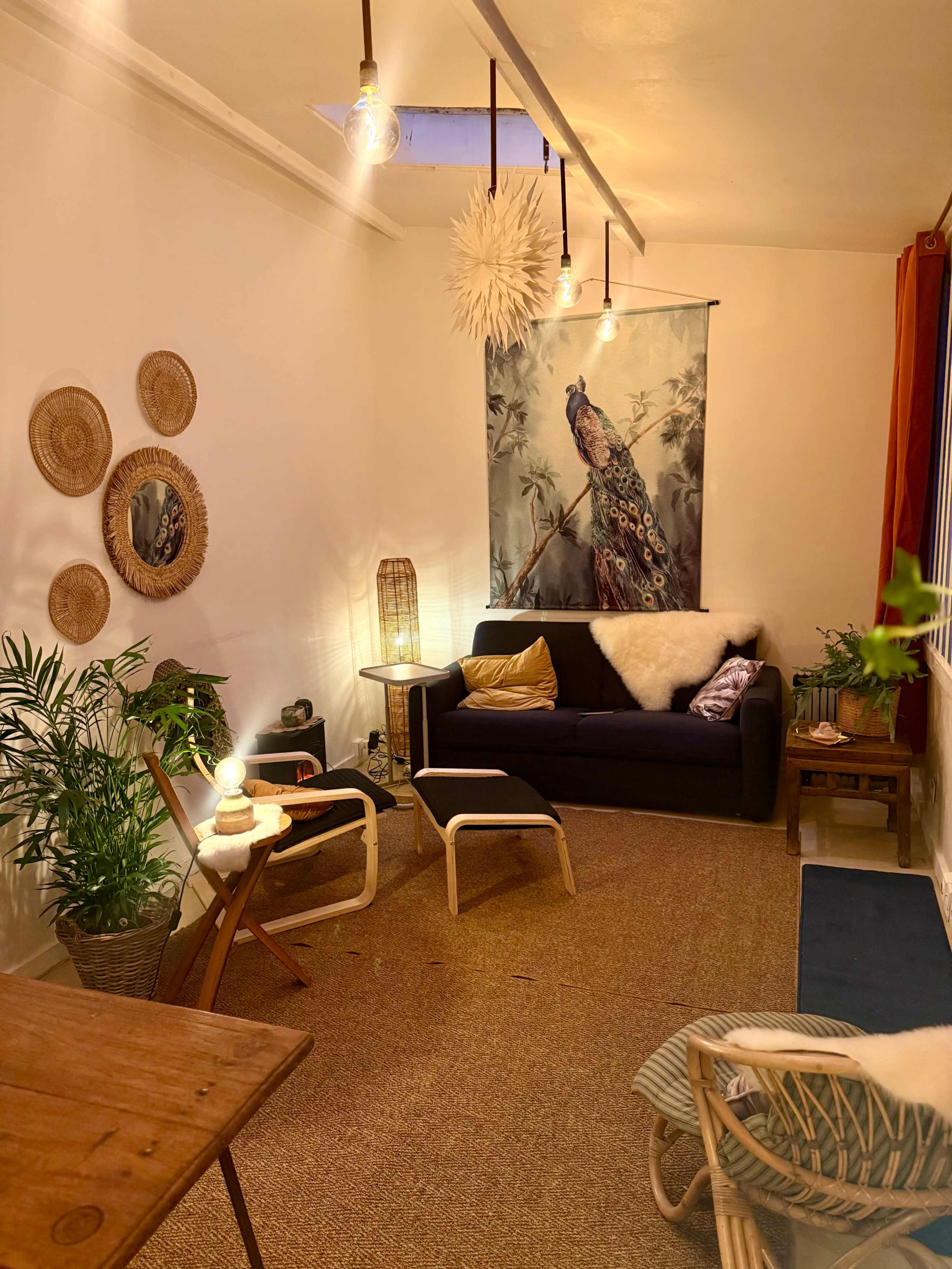 A cozy living room features a dark sofa, white chairs, decorative wall art with a peacock, and plants, illuminated by warm lighting.