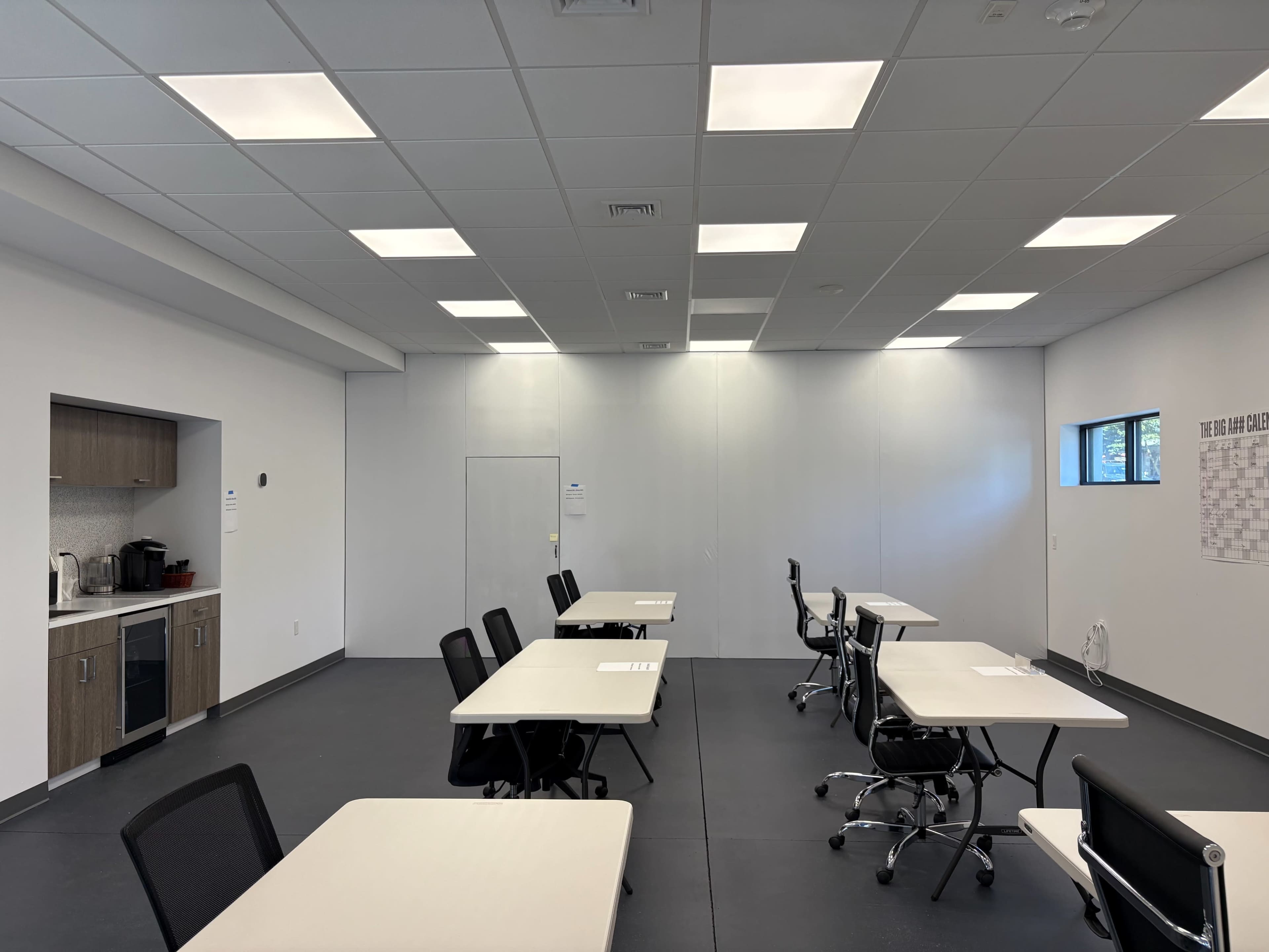 The image shows a spacious, well-lit classroom with several tables and chairs arranged neatly, along with a kitchenette area in the corner.