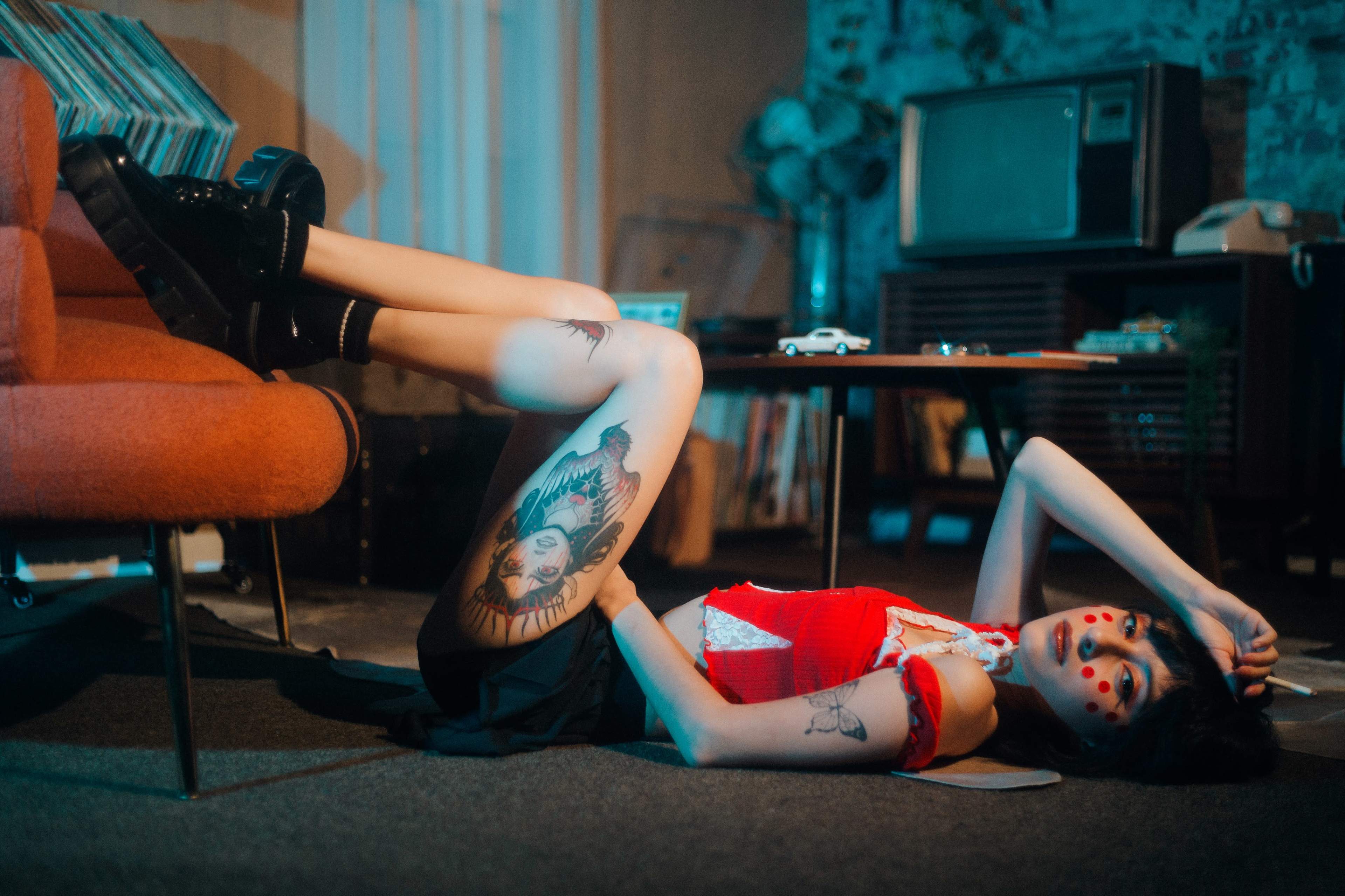 A woman in a red top and black skirt lies on the floor of a retro-styled living room, with her legs propped up on an orange chair.