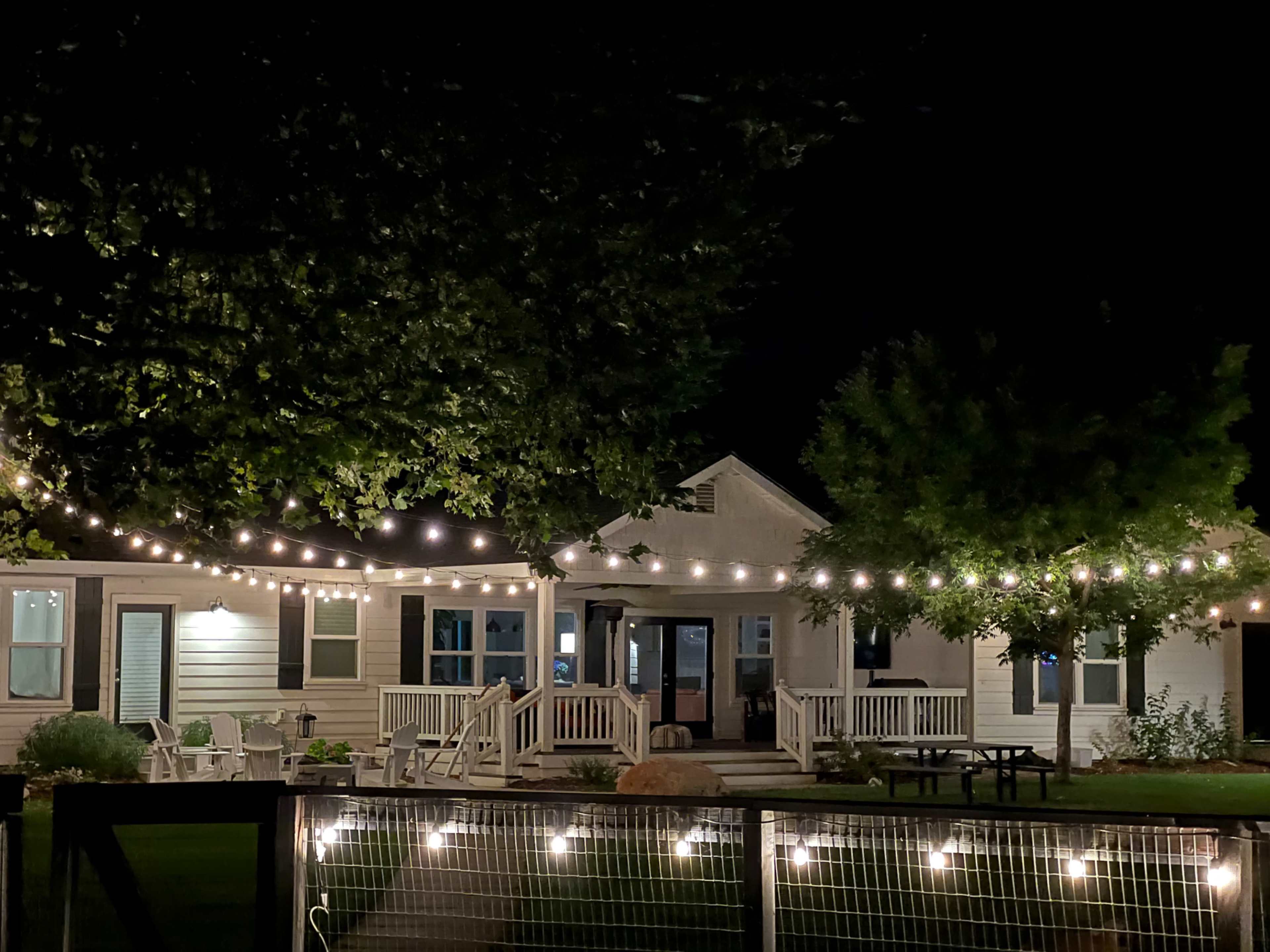 A well-lit house with string lights hangs over a patio area, surrounded by trees at night.