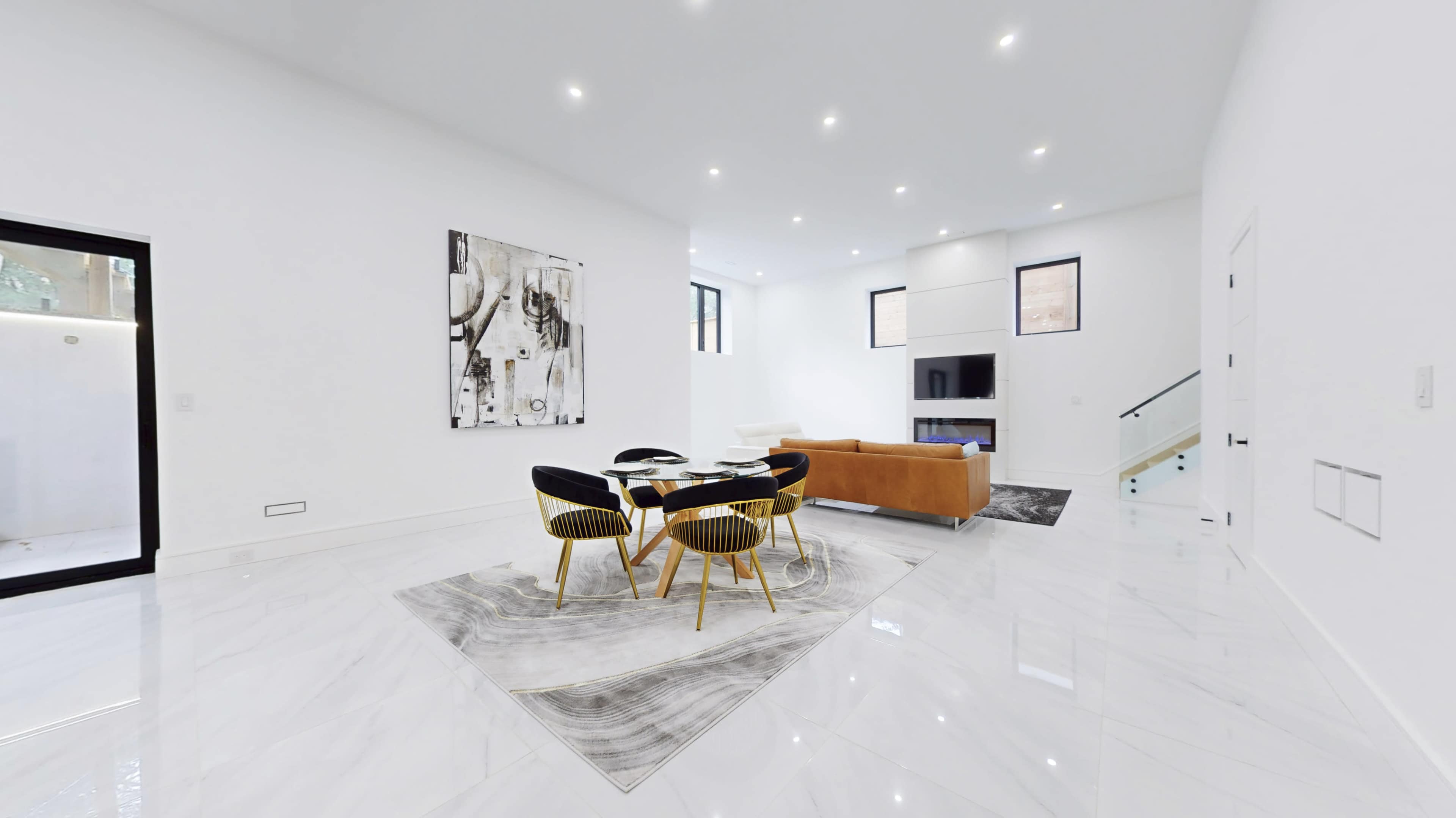 A spacious, modern living area features a round dining table surrounded by black and gold chairs, a large artwork on the wall, and a tan sofa.