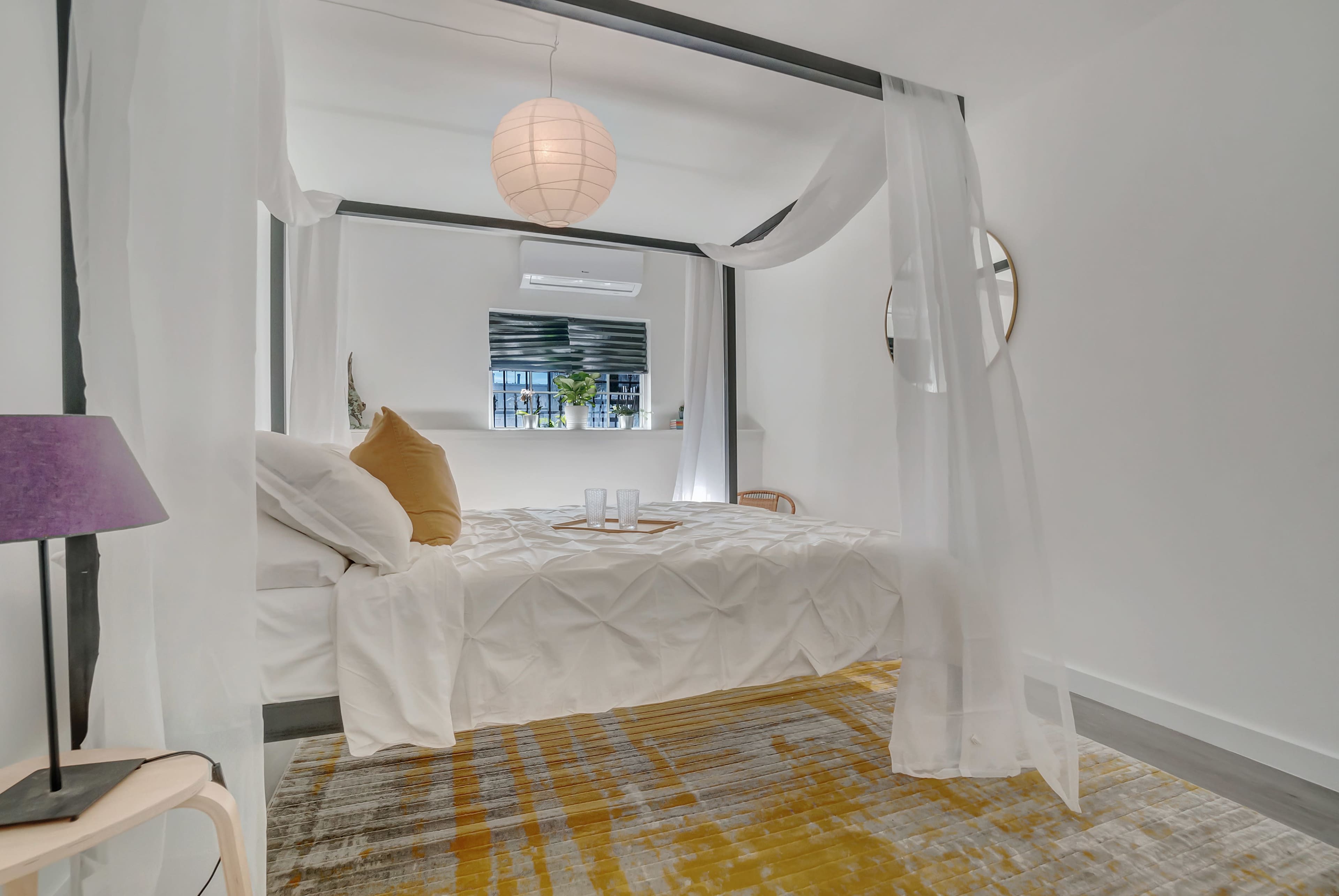 A four-poster bed with sheer drapes is positioned on a textured rug in a minimalist bedroom featuring a round mirror and a small table with drinks.