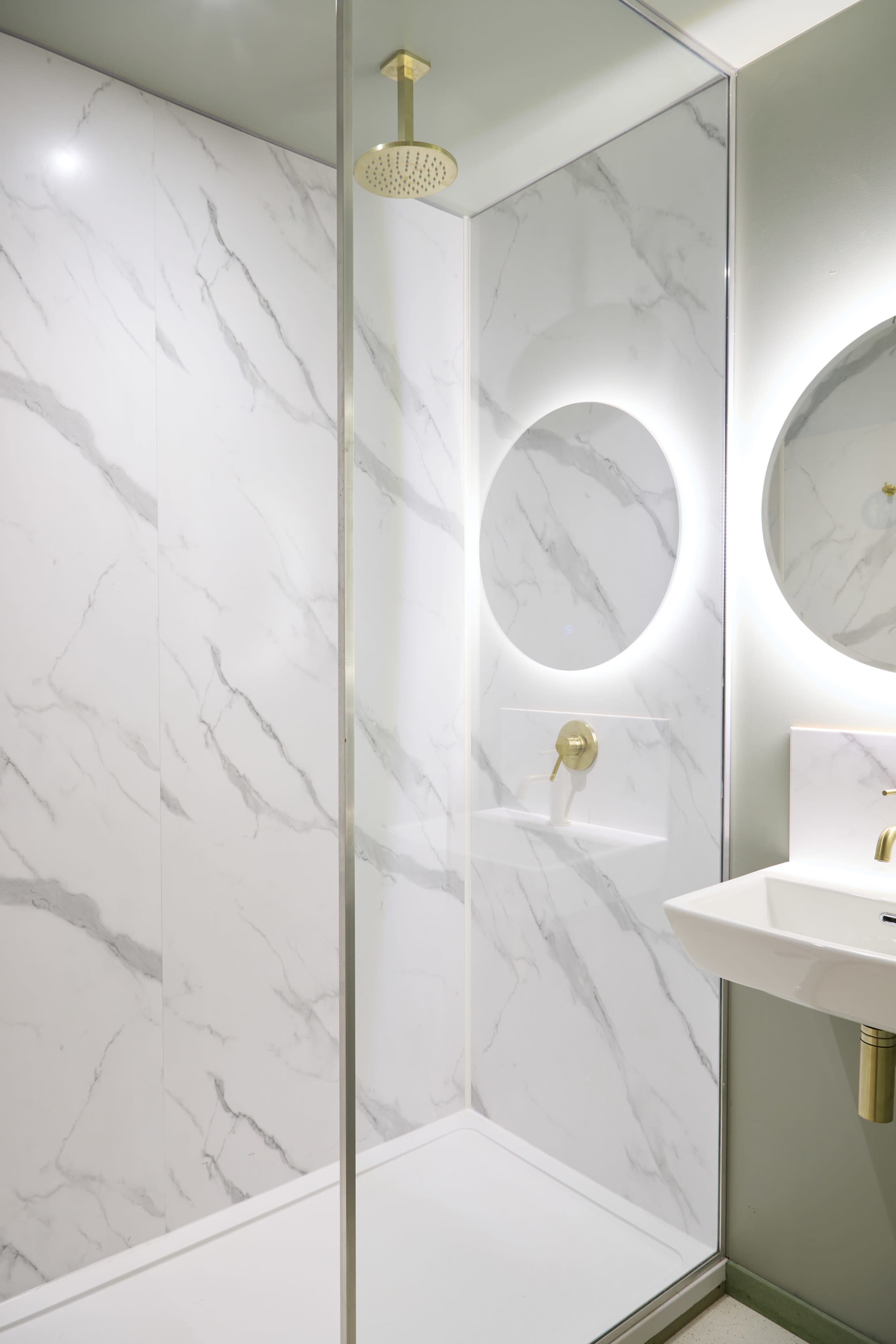 A modern shower stall features marble-patterned walls, a circular mirror with backlighting, and a minimalist sink.
