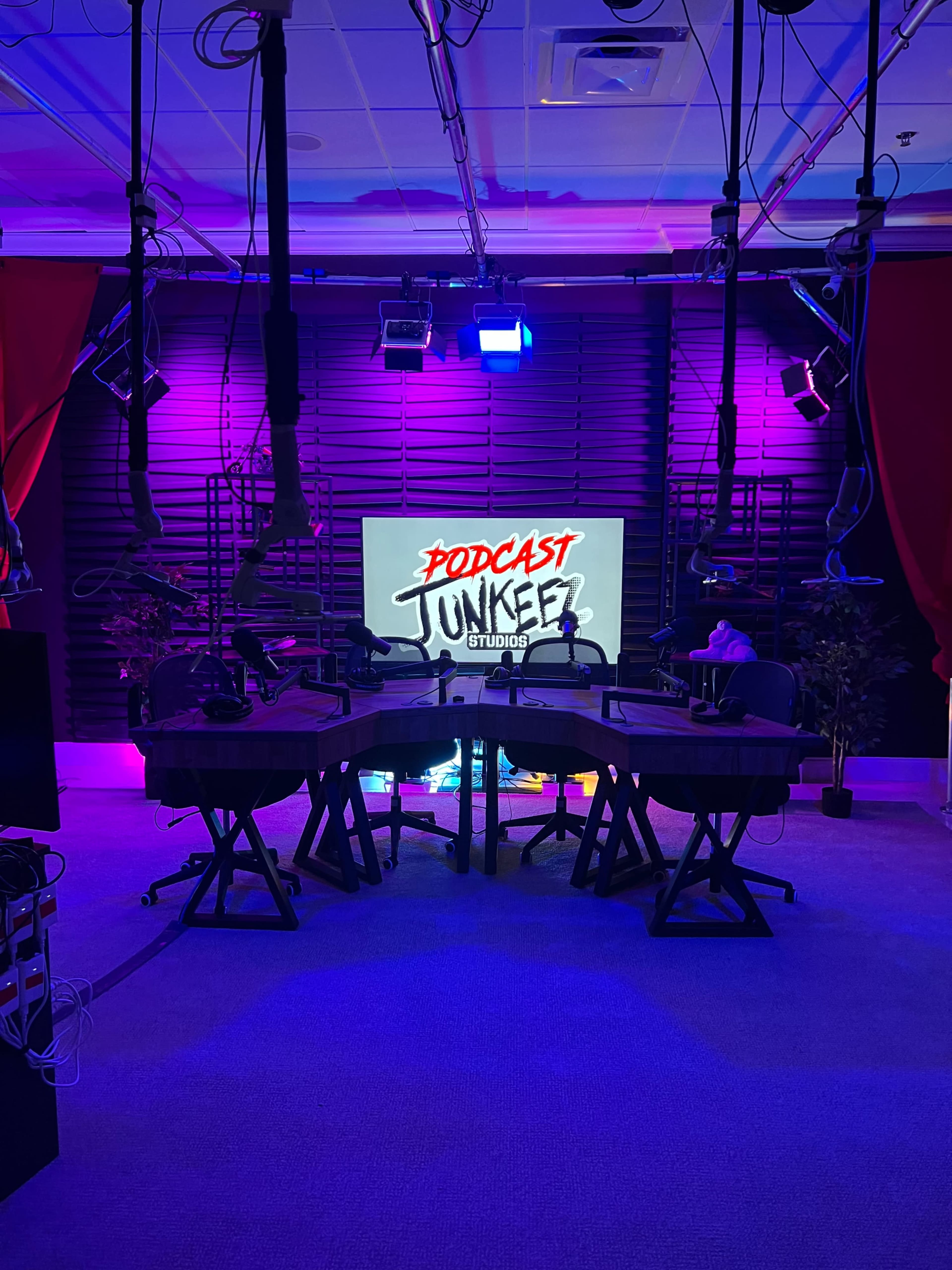 The image shows a podcast studio featuring a central table surrounded by microphones, with a large screen displaying "PODCAST JUNKIEZ STUDIOS" in colorful text against a backdrop of purple lighting.