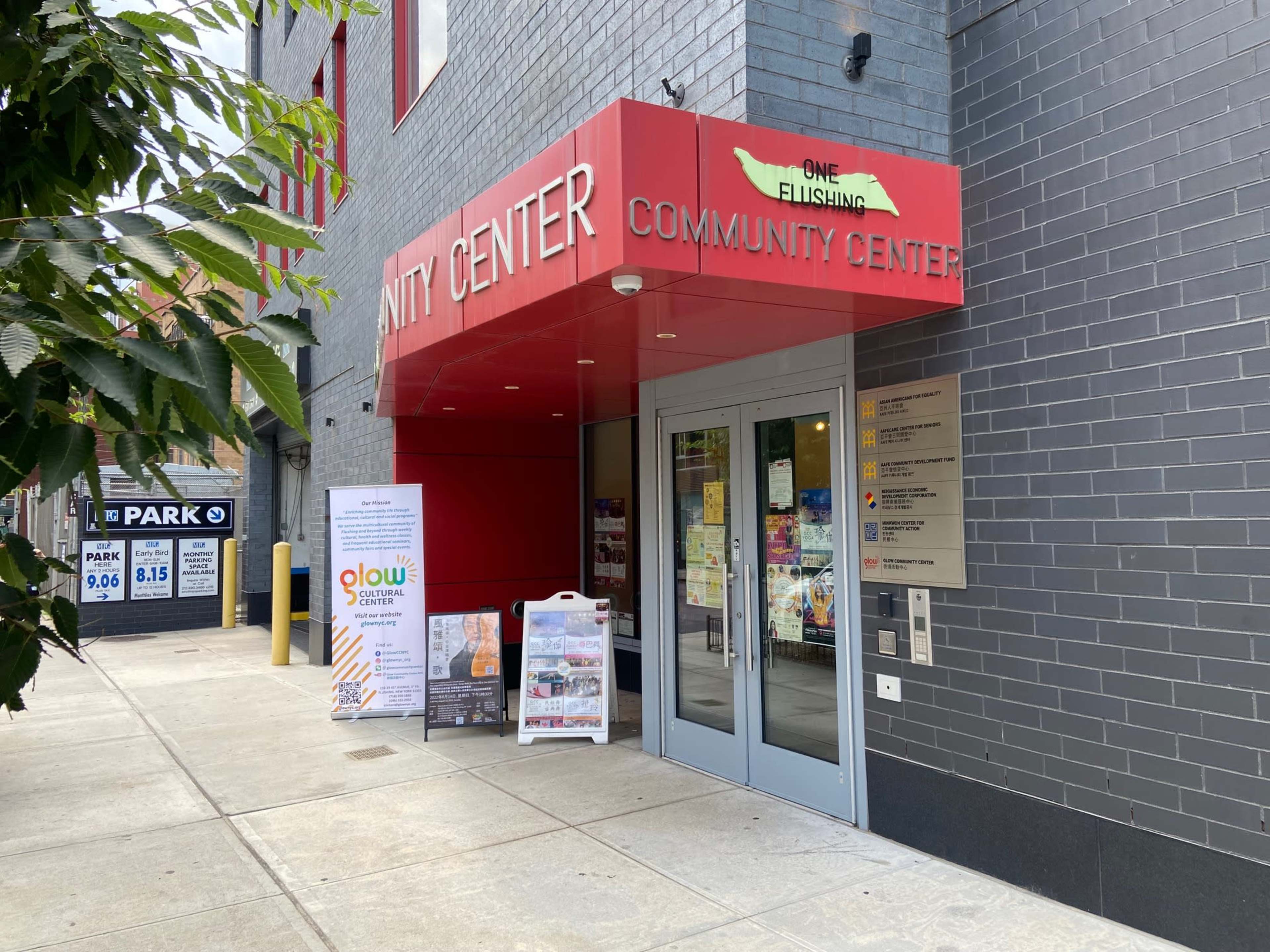 The entrance to the One Flushing Community Center features red signage and a sidewalk display board with promotional materials.