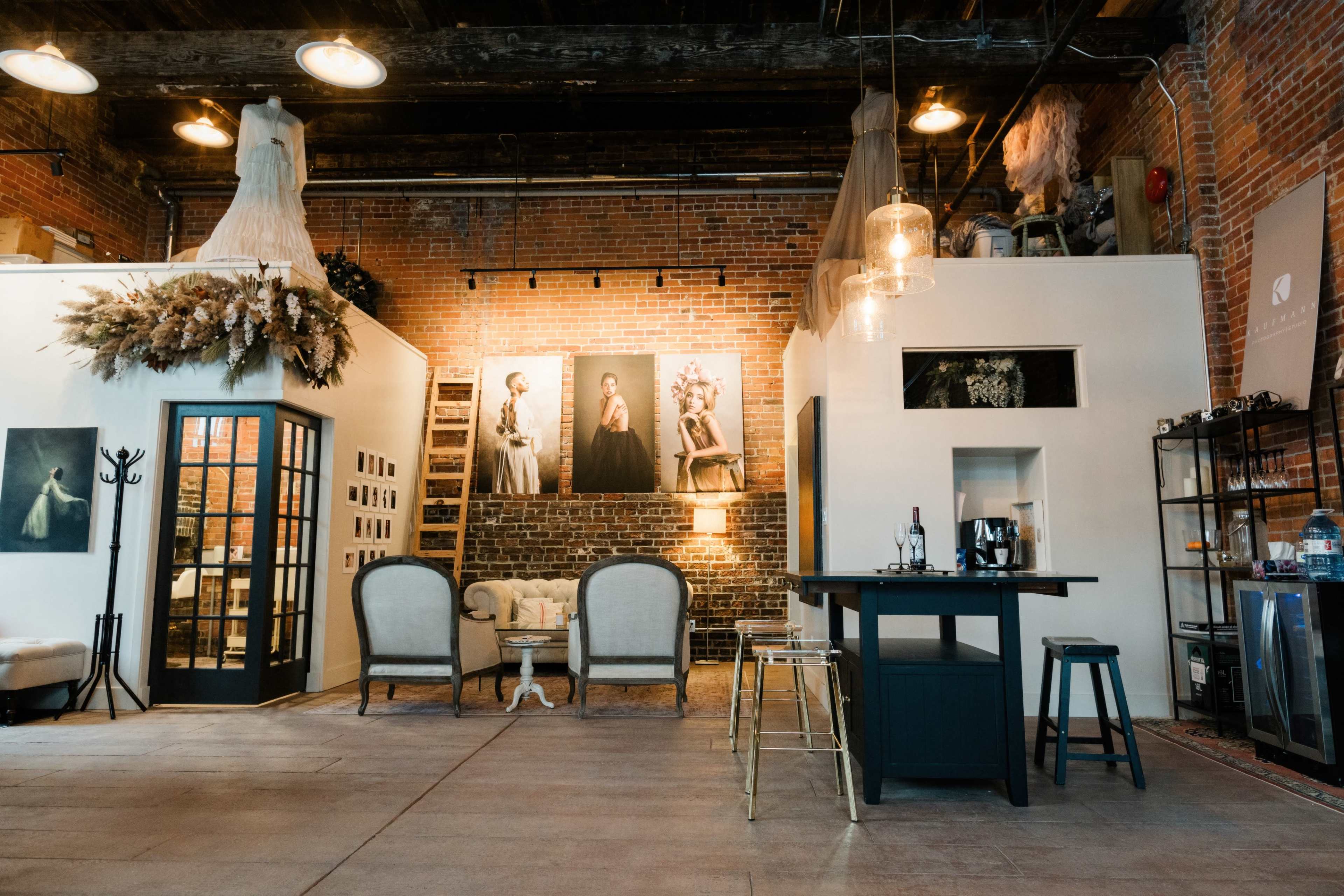 A spacious loft-style interior with exposed brick walls, a seating area, a bar-style table, and artistic photographs displayed on the walls.