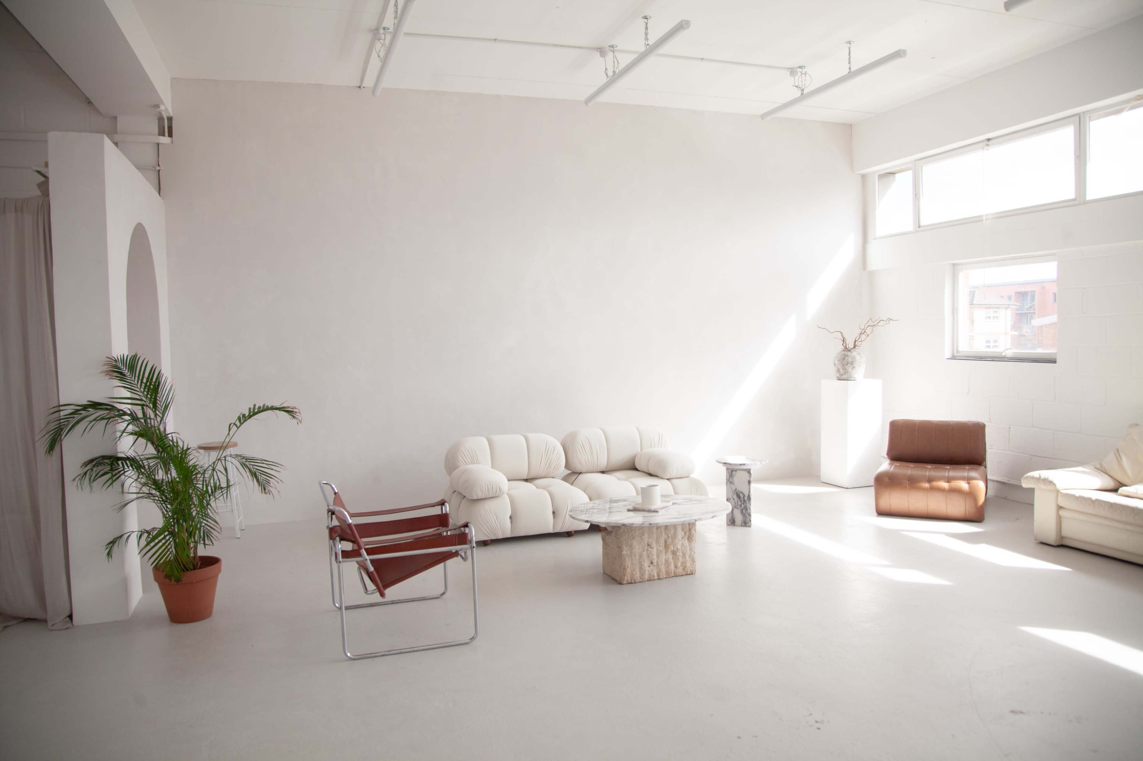 The image shows a minimalist interior space featuring a white sofa, a brown armchair, a wooden coffee table, and a potted plant, with large windows allowing natural light to illuminate the room.