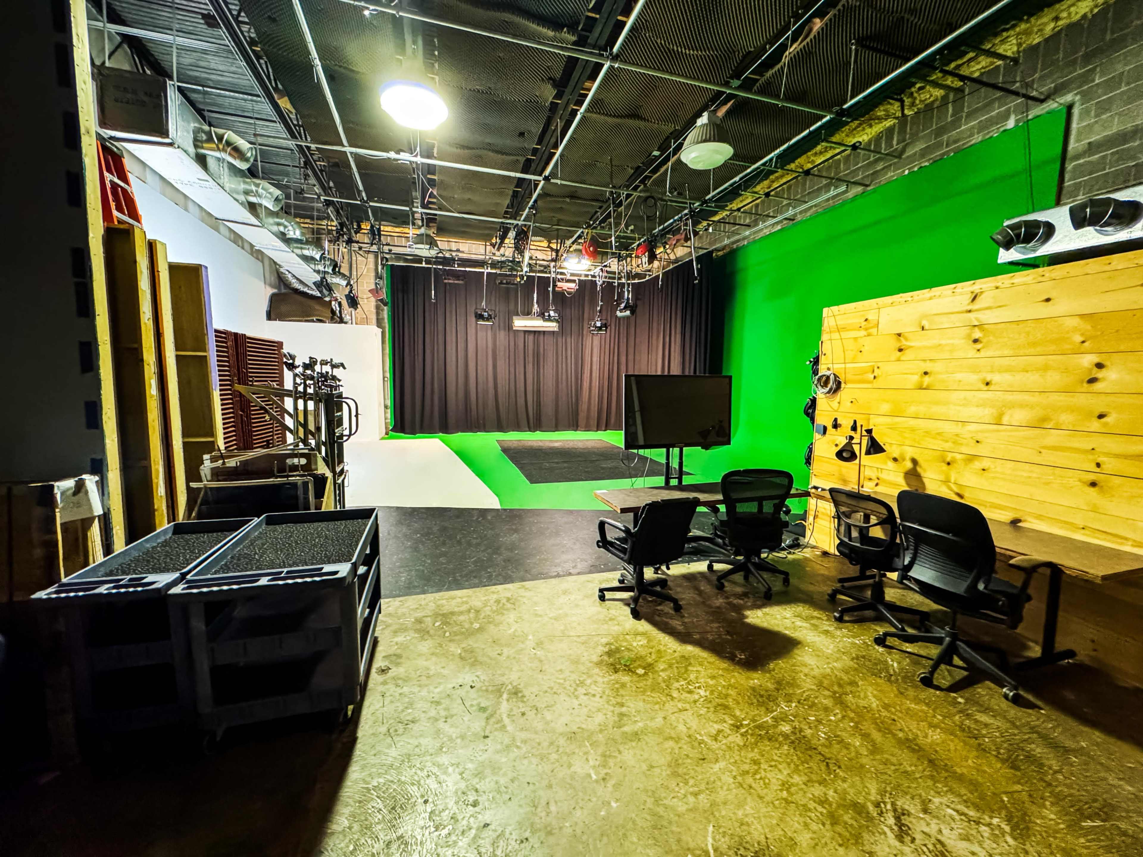 The image shows a recording studio with a green screen backdrop, black curtains, and various equipment, including chairs and a monitor.