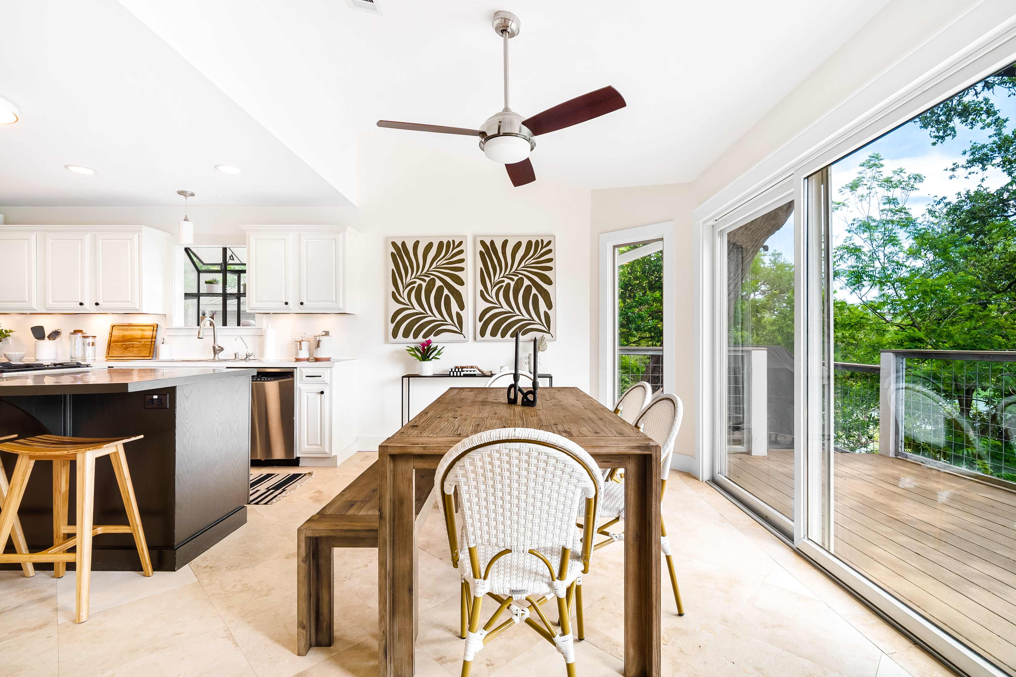 A modern kitchen and dining area features a wooden dining table surrounded by chairs, a ceiling fan, and sliding glass doors leading to a deck.