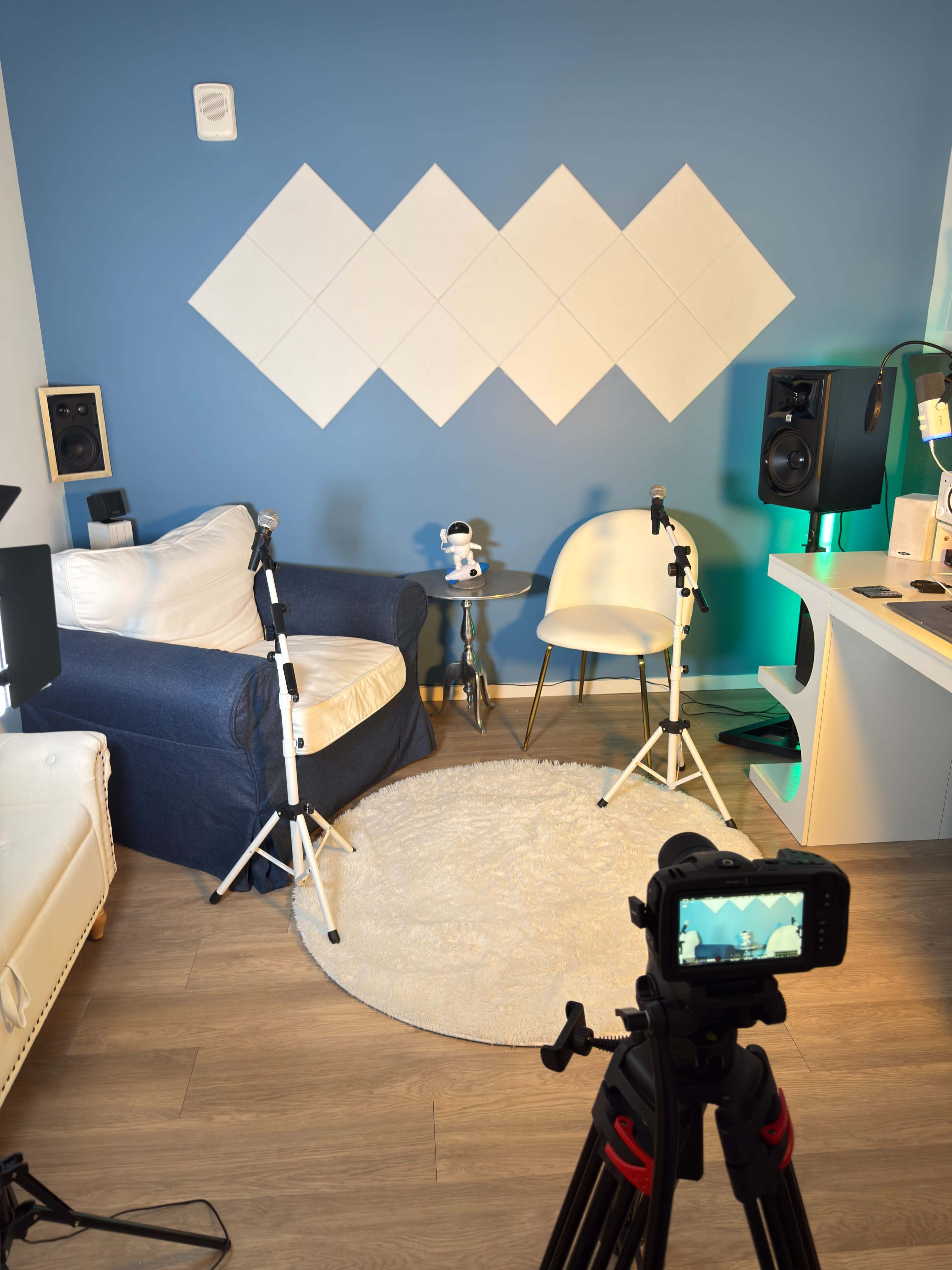 The image shows a cozy room setup featuring a blue wall with white diamond shapes, a white rug on a wooden floor, a blue armchair, a small table, and camera tripods positioned in front of a camera on a tripod.