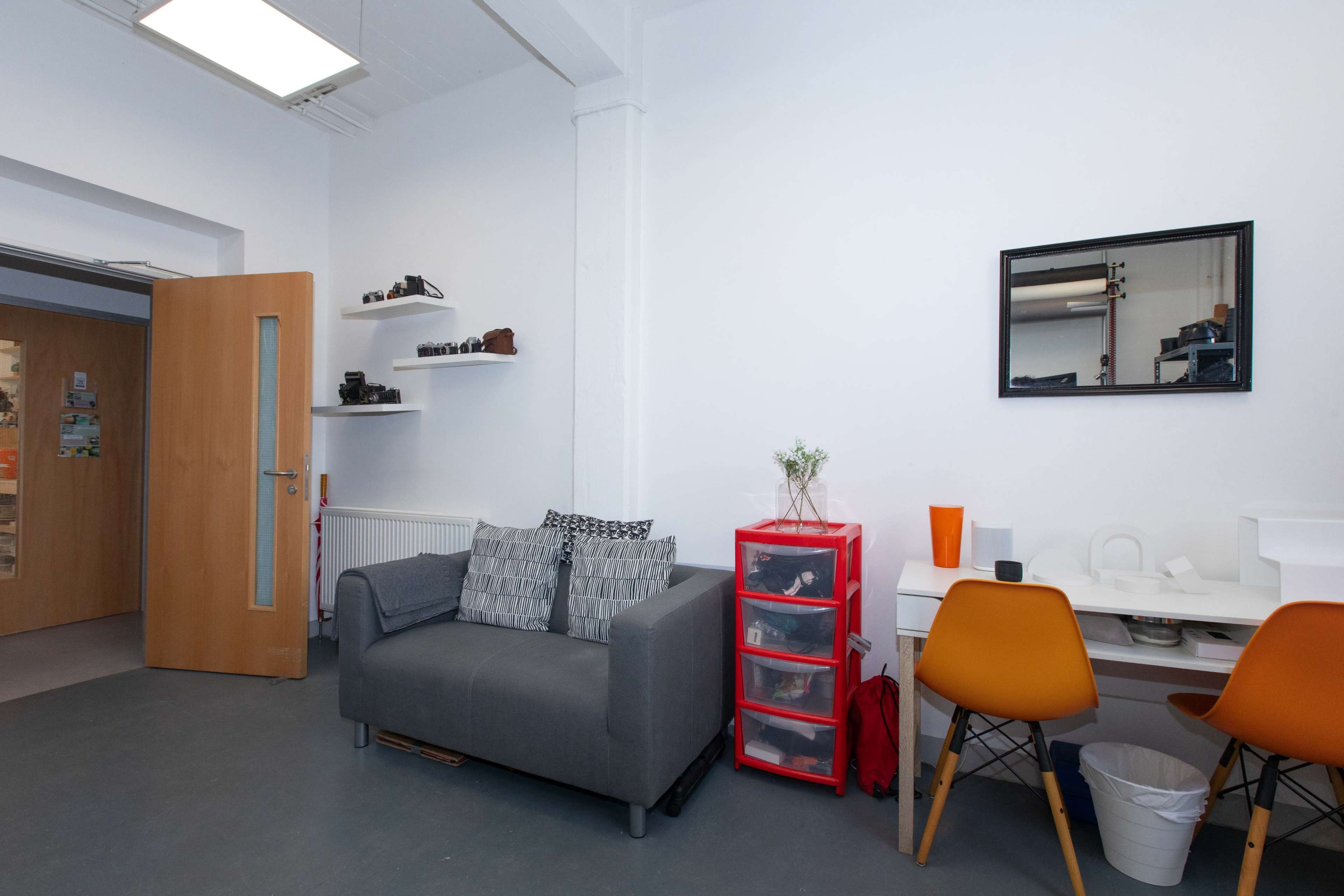 A small, minimalistic workspace featuring a gray sofa, a red storage unit, a white desk with an orange vase, and a mirror on the wall, along with wooden doors leading to another room.