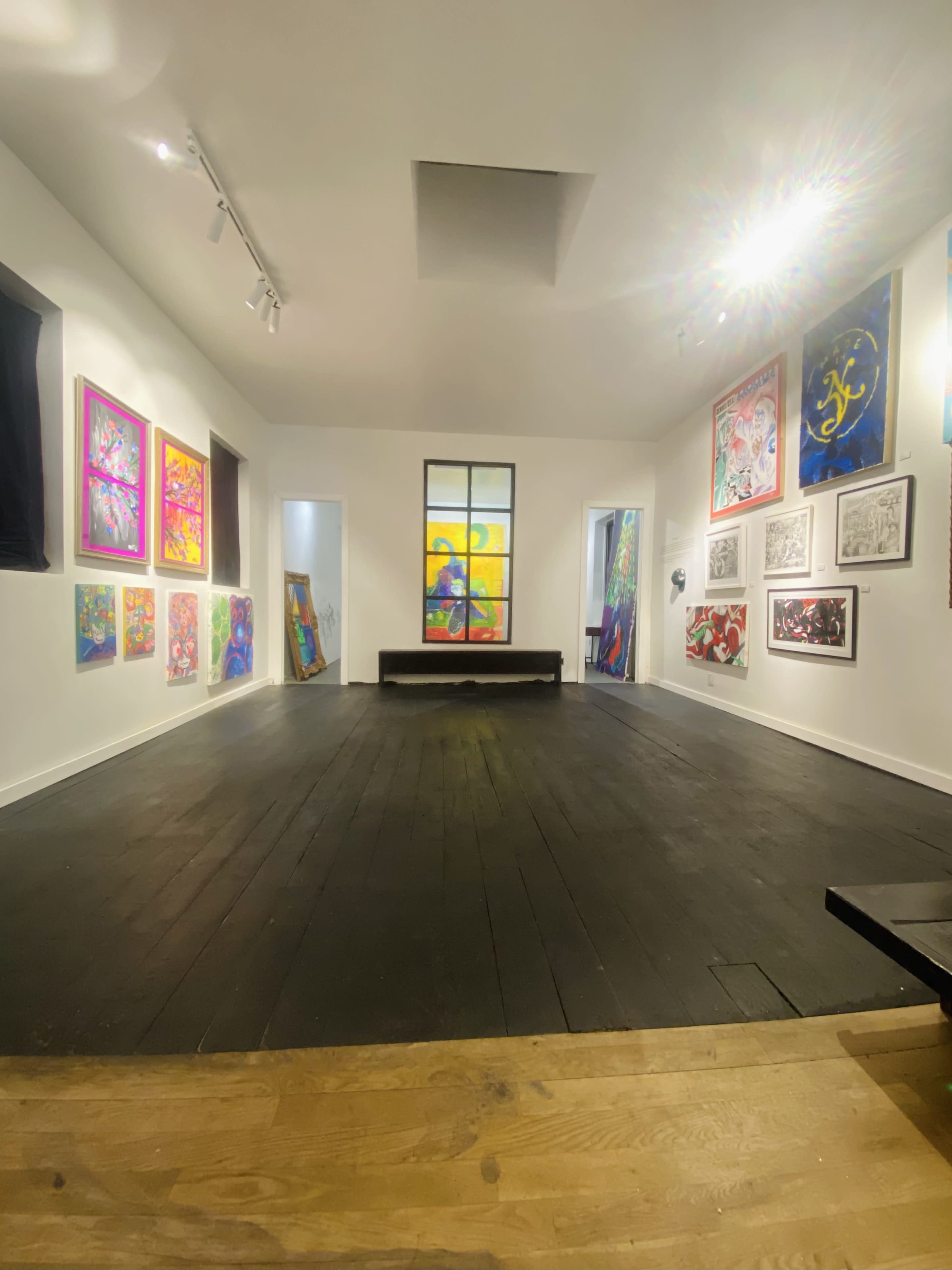The image shows an art gallery with colorful paintings displayed on the white walls and a wooden floor.