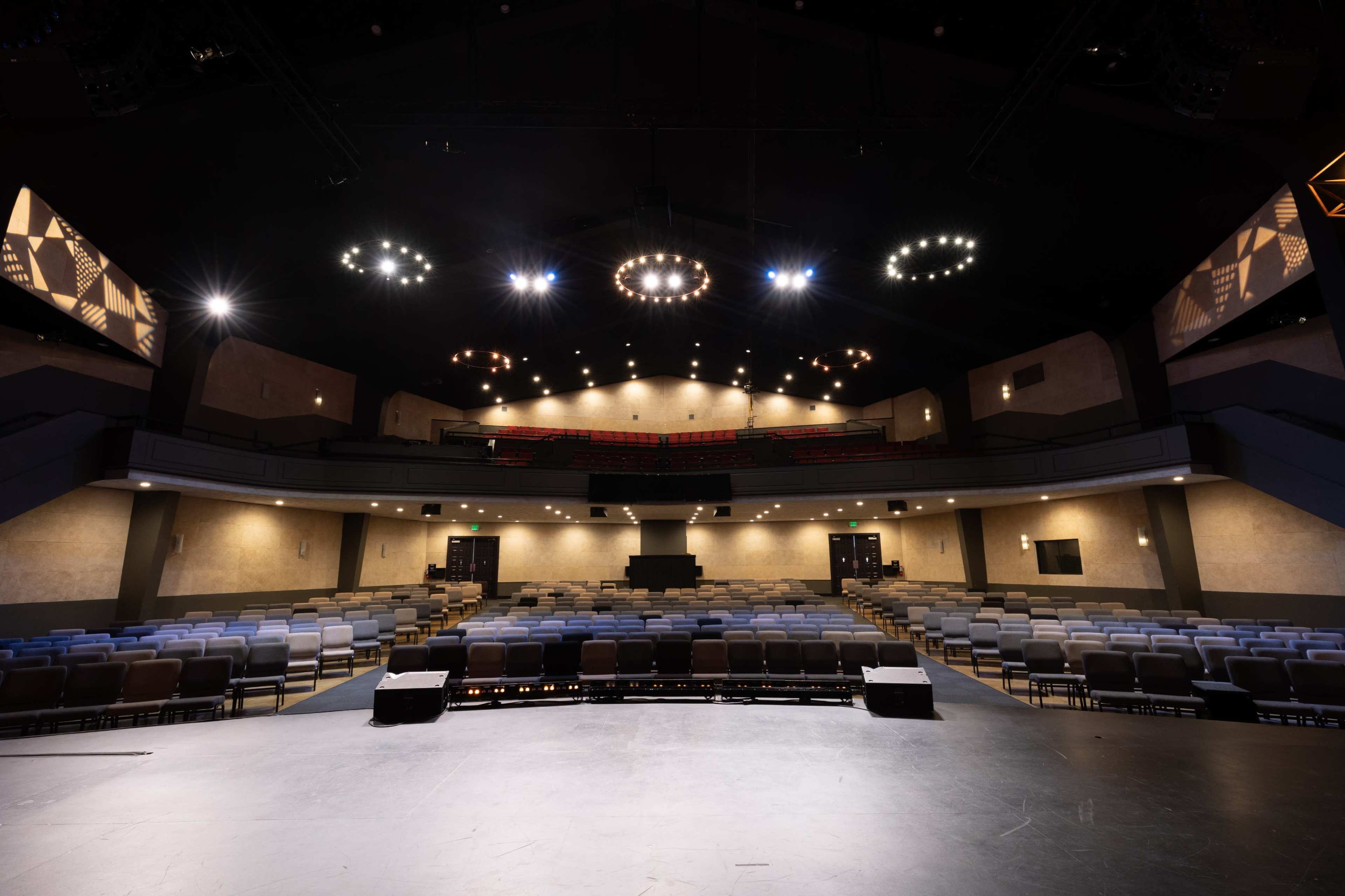 The image shows the interior of a theater with tiered seating and a stage illuminated by overhead lights.