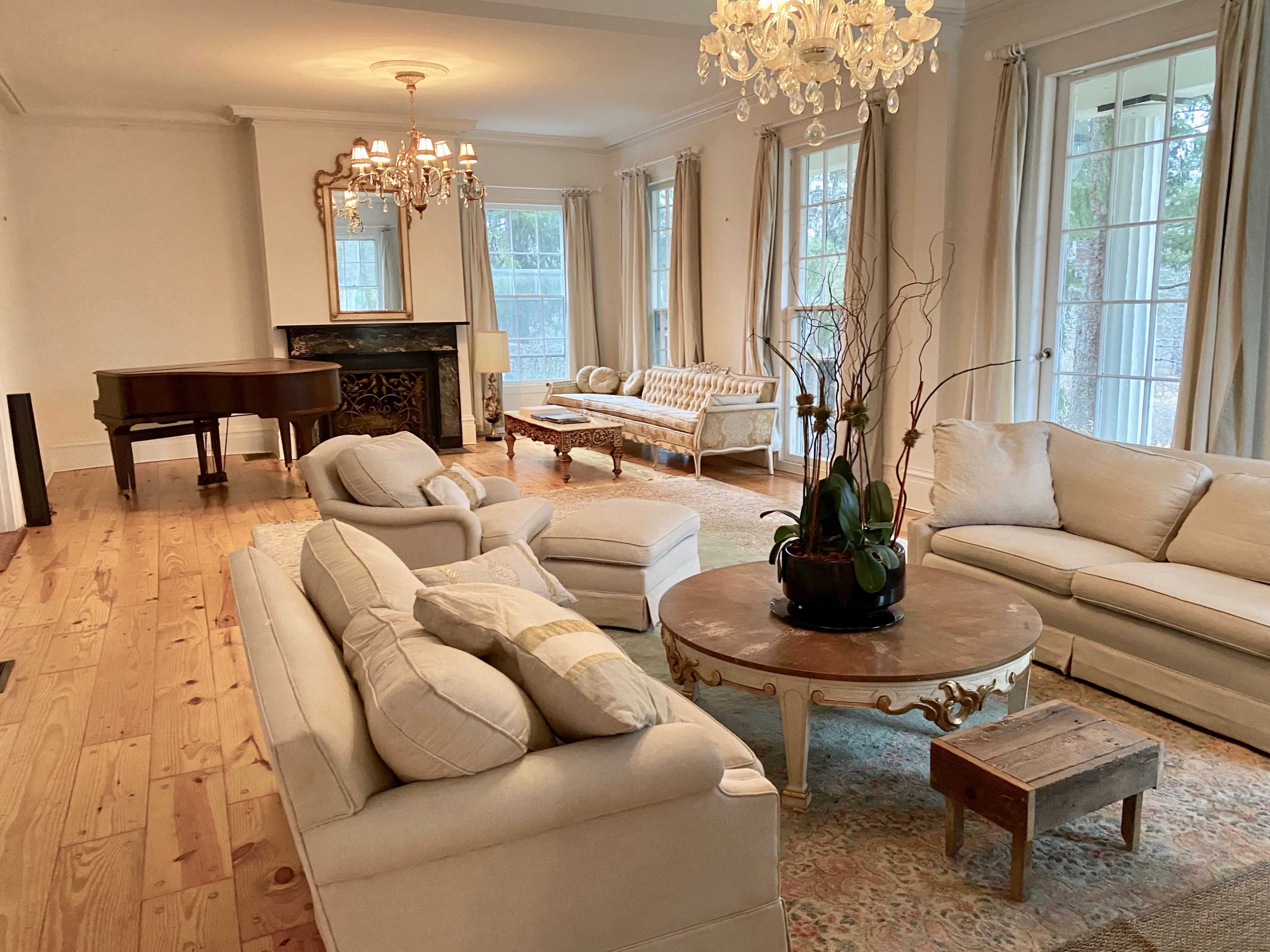 The spacious living room features a grand piano, a chandelier, and various comfortable seating arrangements around a round table.