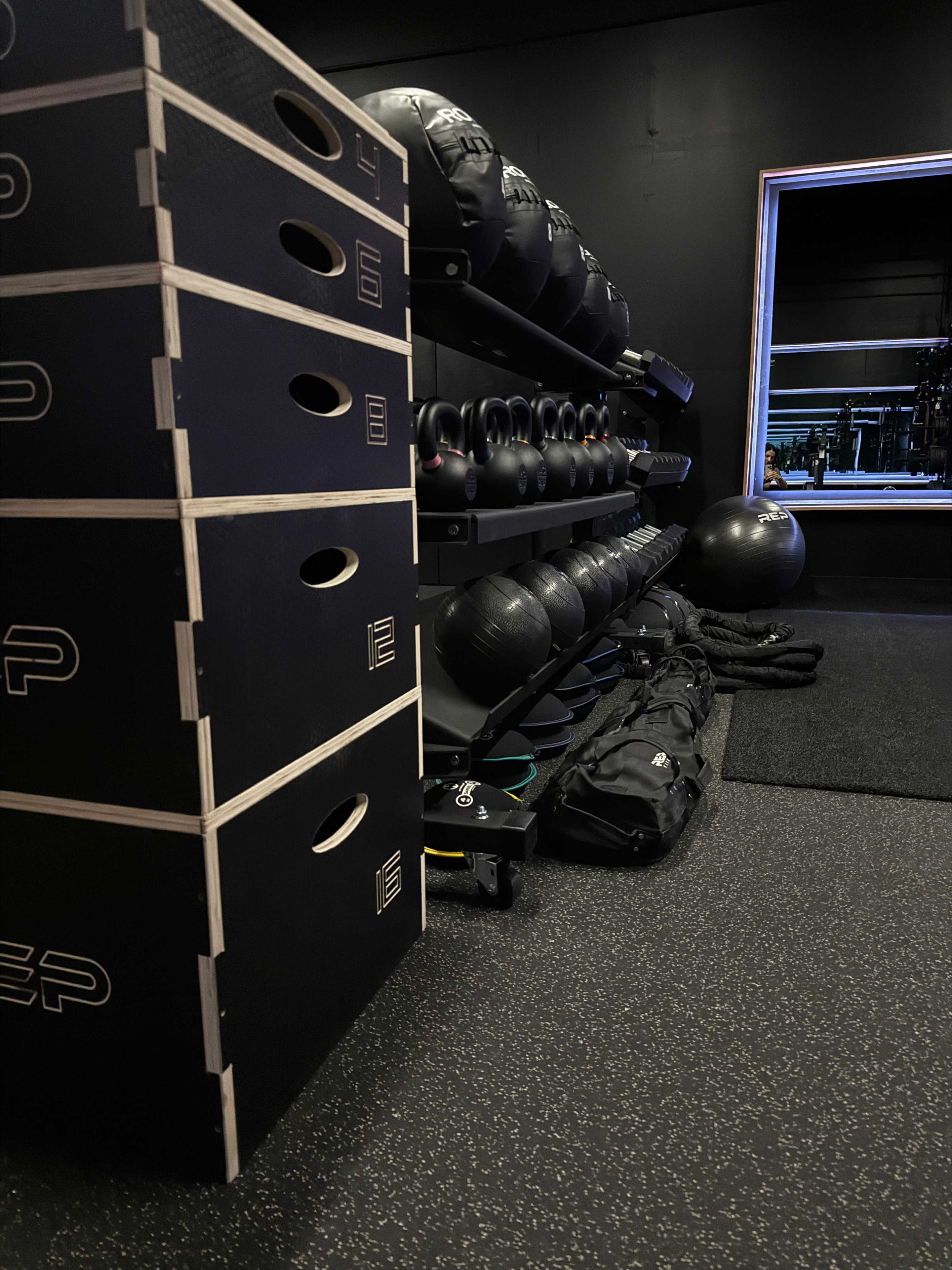 The image shows a corner of a gym featuring stacked plyometric boxes, exercise balls, and fitness bags against a dark wall.