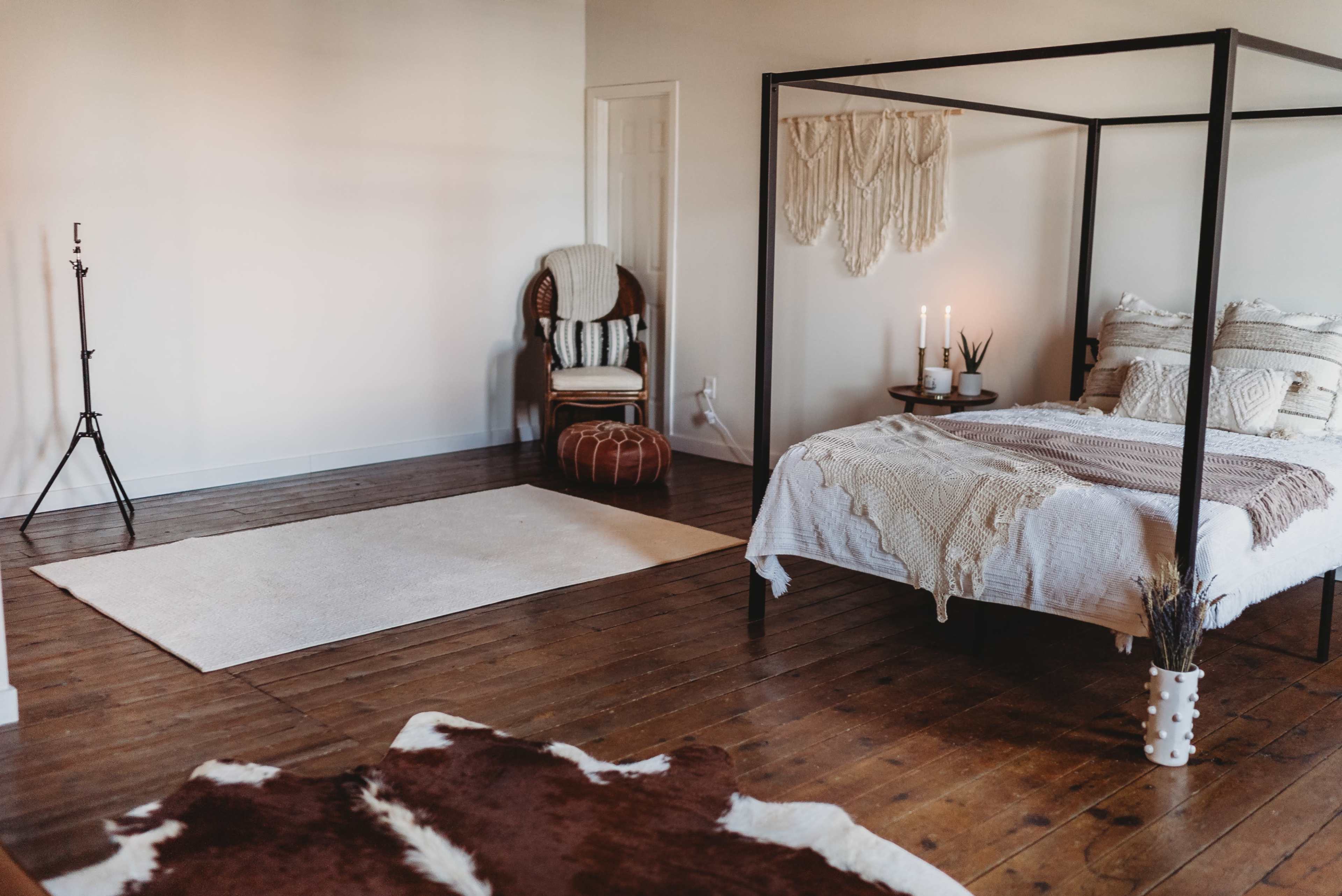 The image shows a minimalistic bedroom featuring a four-poster bed, a woven wall hanging, a chair, and a floor rug on wooden flooring.