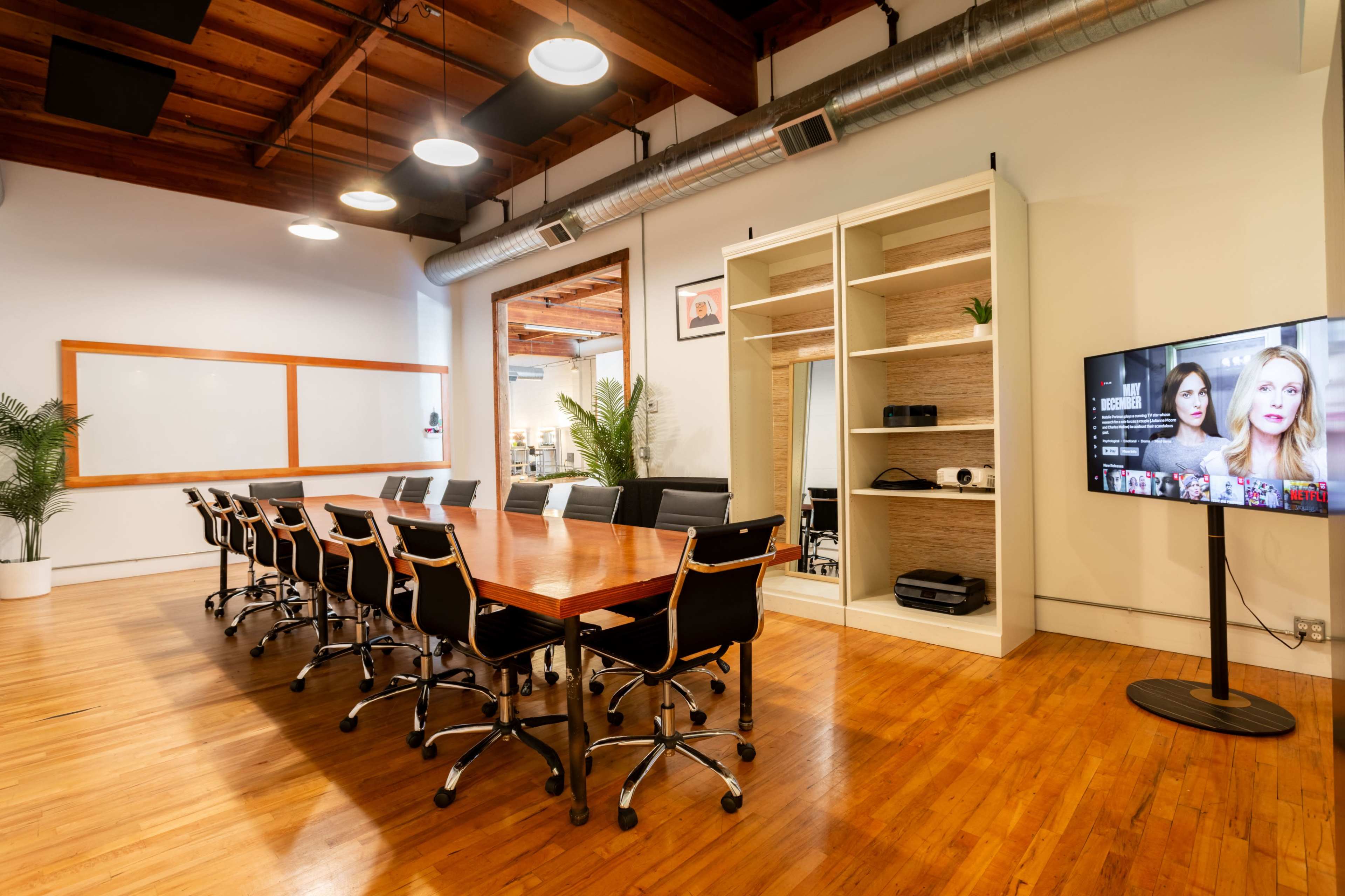 A modern conference room features a long wooden table surrounded by black chairs, with a television and a shelving unit against one wall.