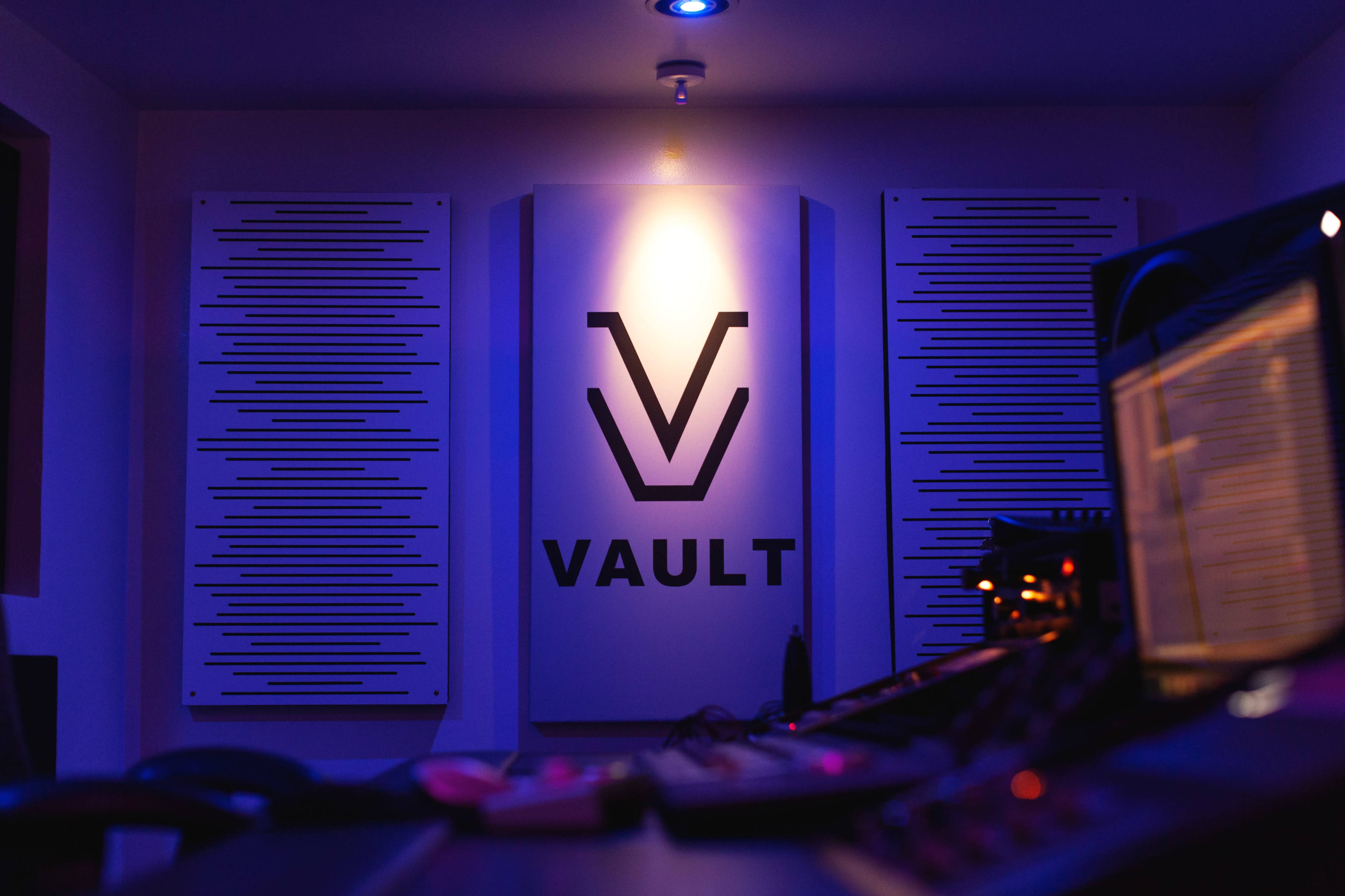 A dimly lit recording studio with a "VAULT" logo prominently displayed on the wall and audio equipment in the foreground.