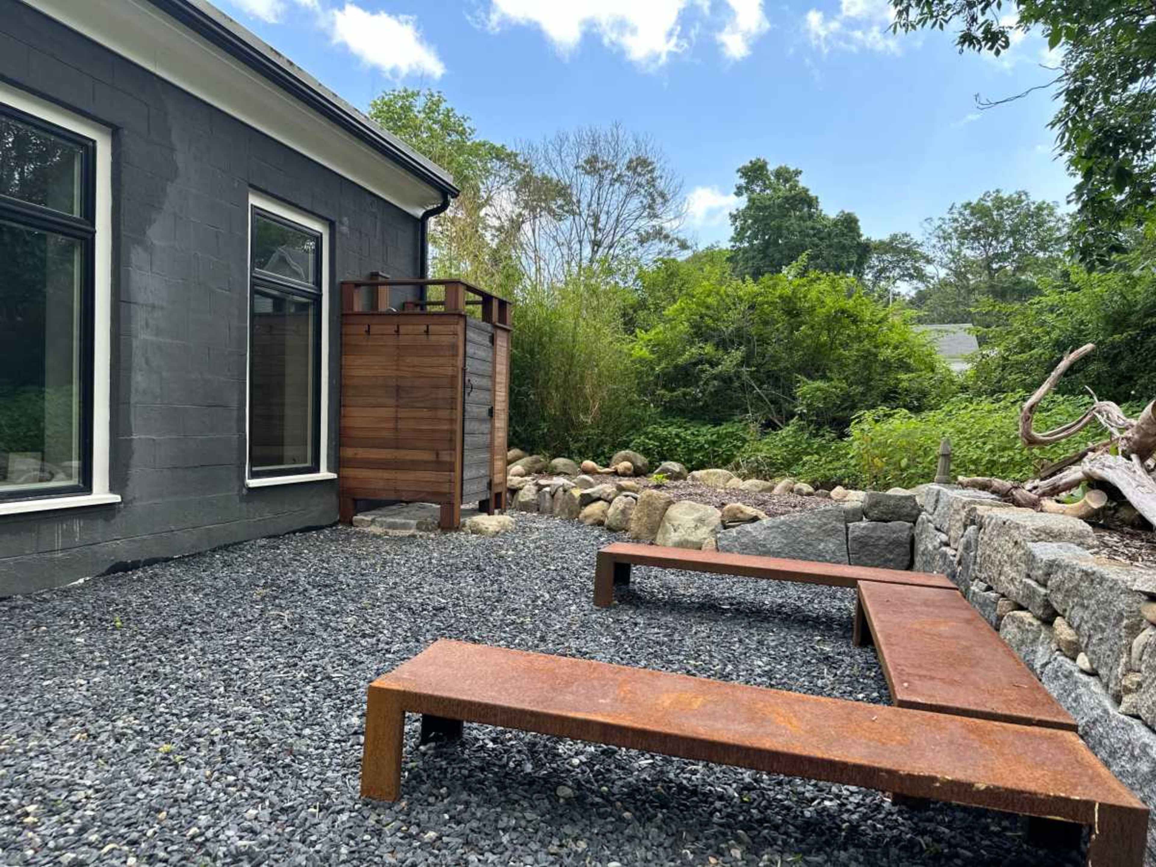 A gravel patio with two rusted metal benches adjacent to a grey house and surrounded by greenery and rocks.
