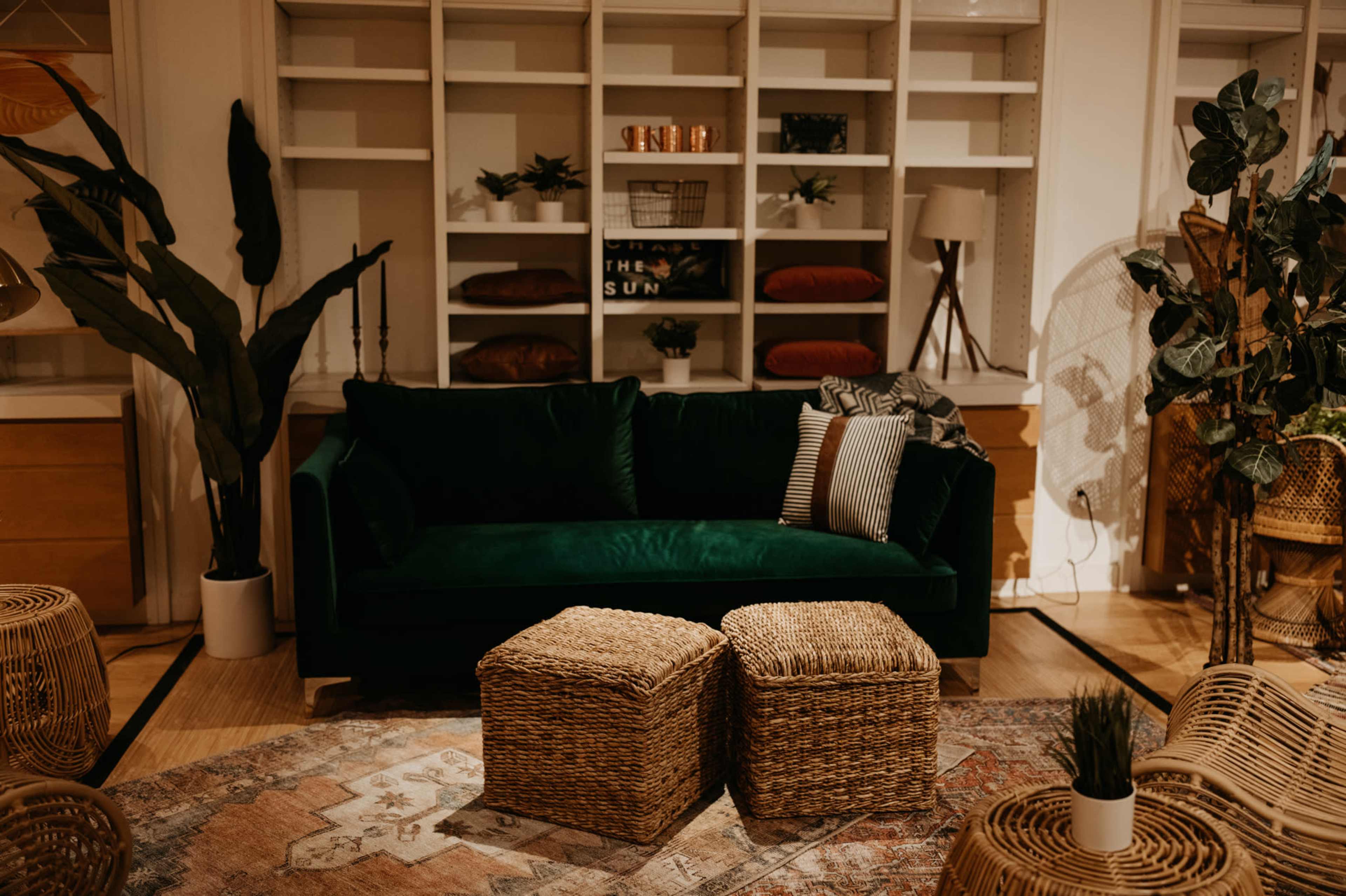 A cozy living room setup featuring a green velvet sofa, two wicker ottomans, and various plants arranged around a bookshelf.