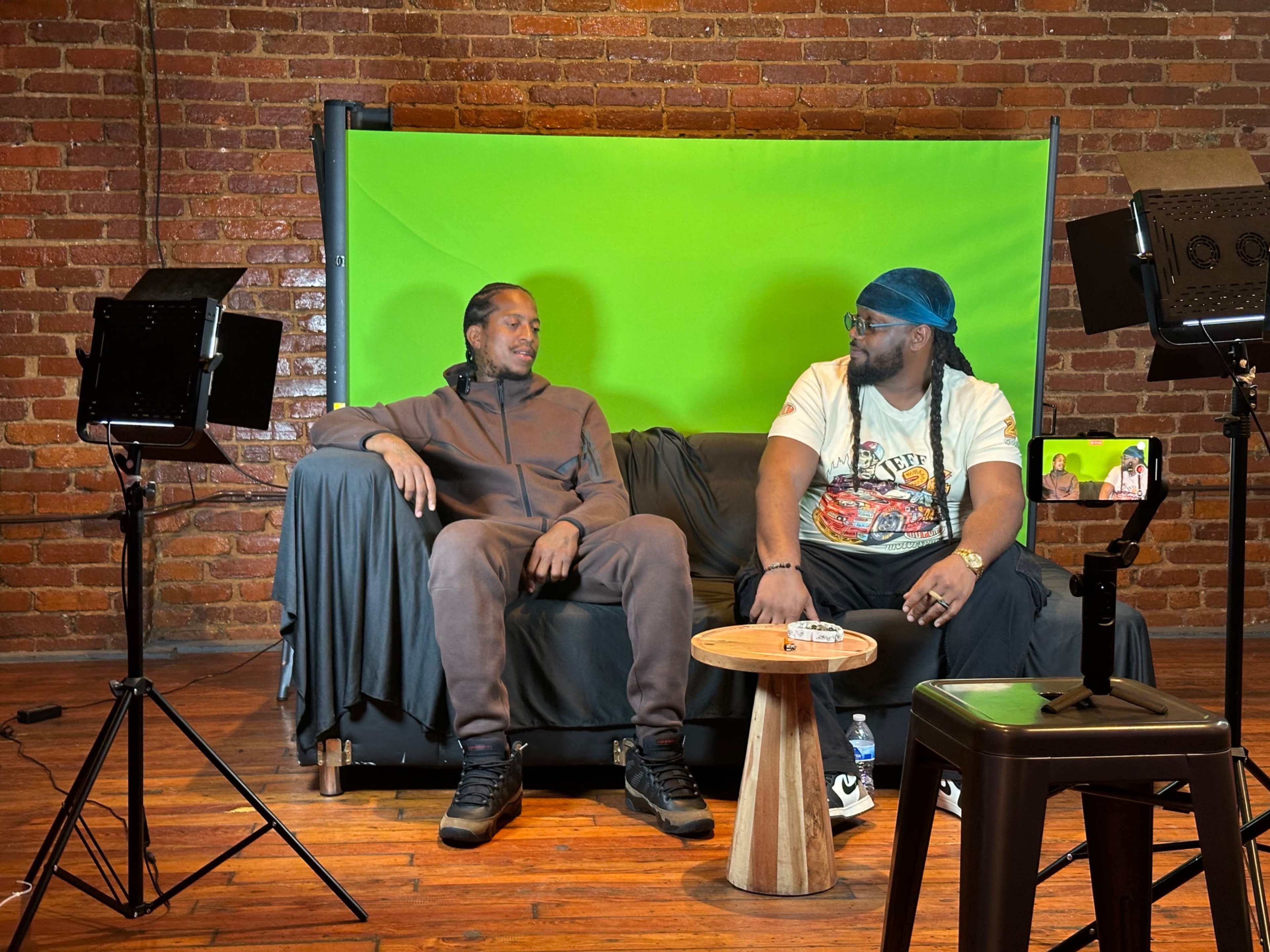 Two men sit on a couch in front of a green screen, with lighting equipment and a small table placed between them.