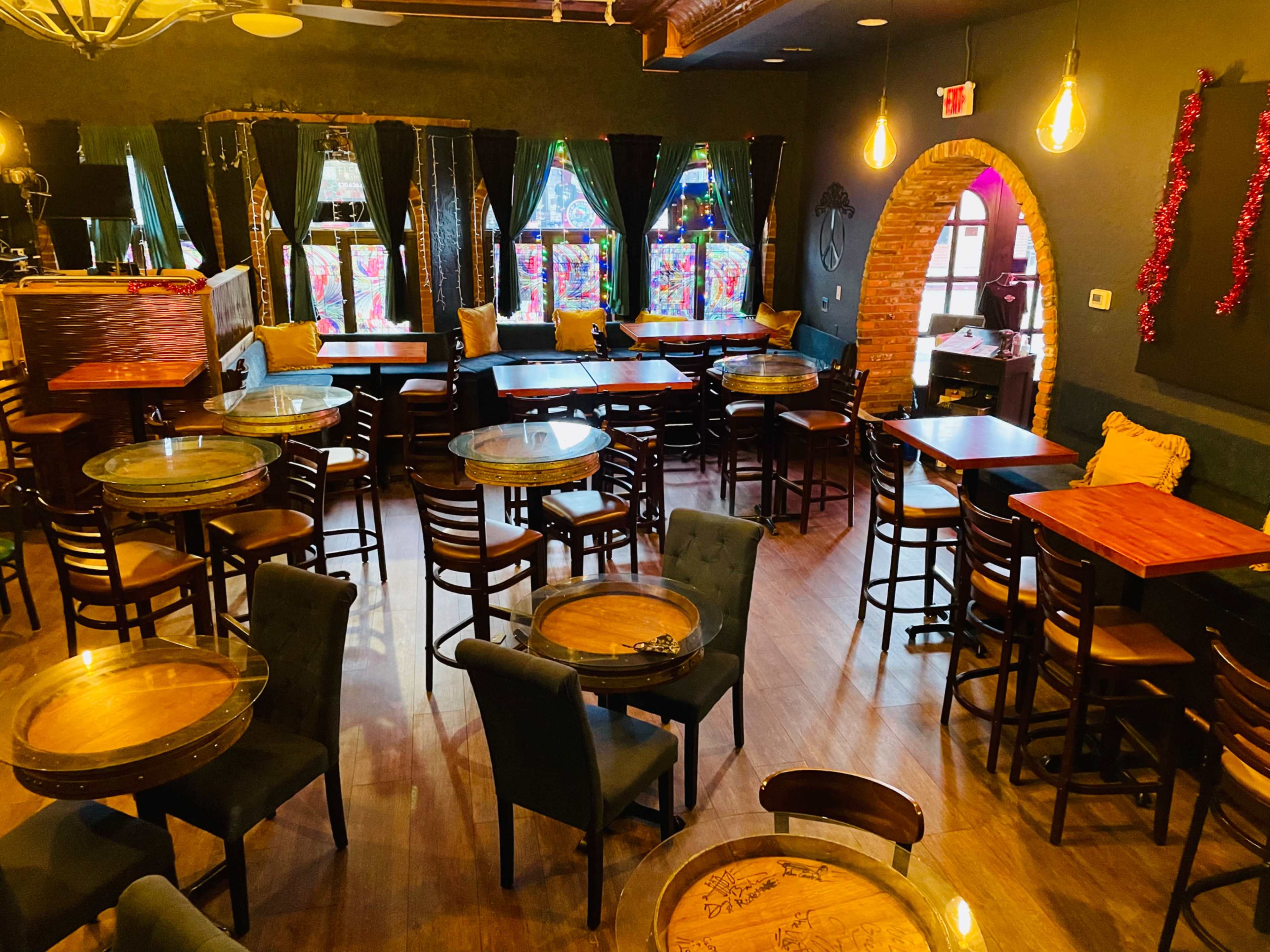 A cozy interior of a café or bar with wooden tables and chairs, large windows featuring colorful stained glass, and dim lighting.
