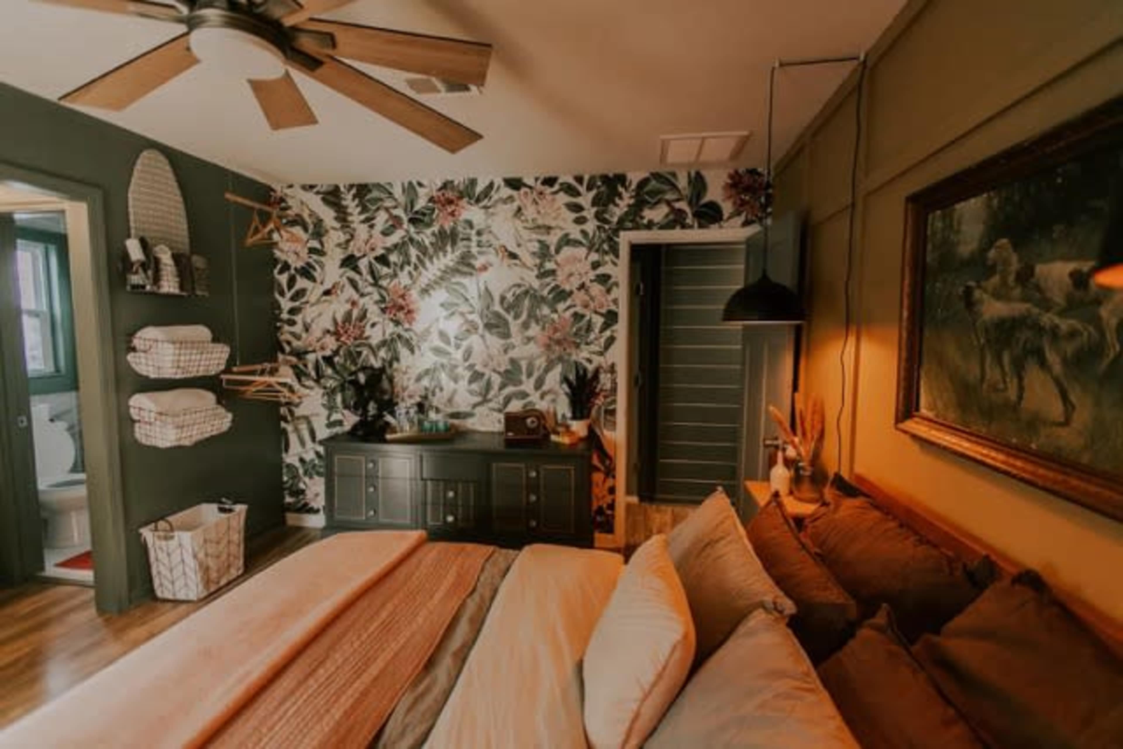 A bedroom features a floral accent wall, a wooden dresser, and a bed with layered linens, alongside a door leading to an adjoining bathroom.
