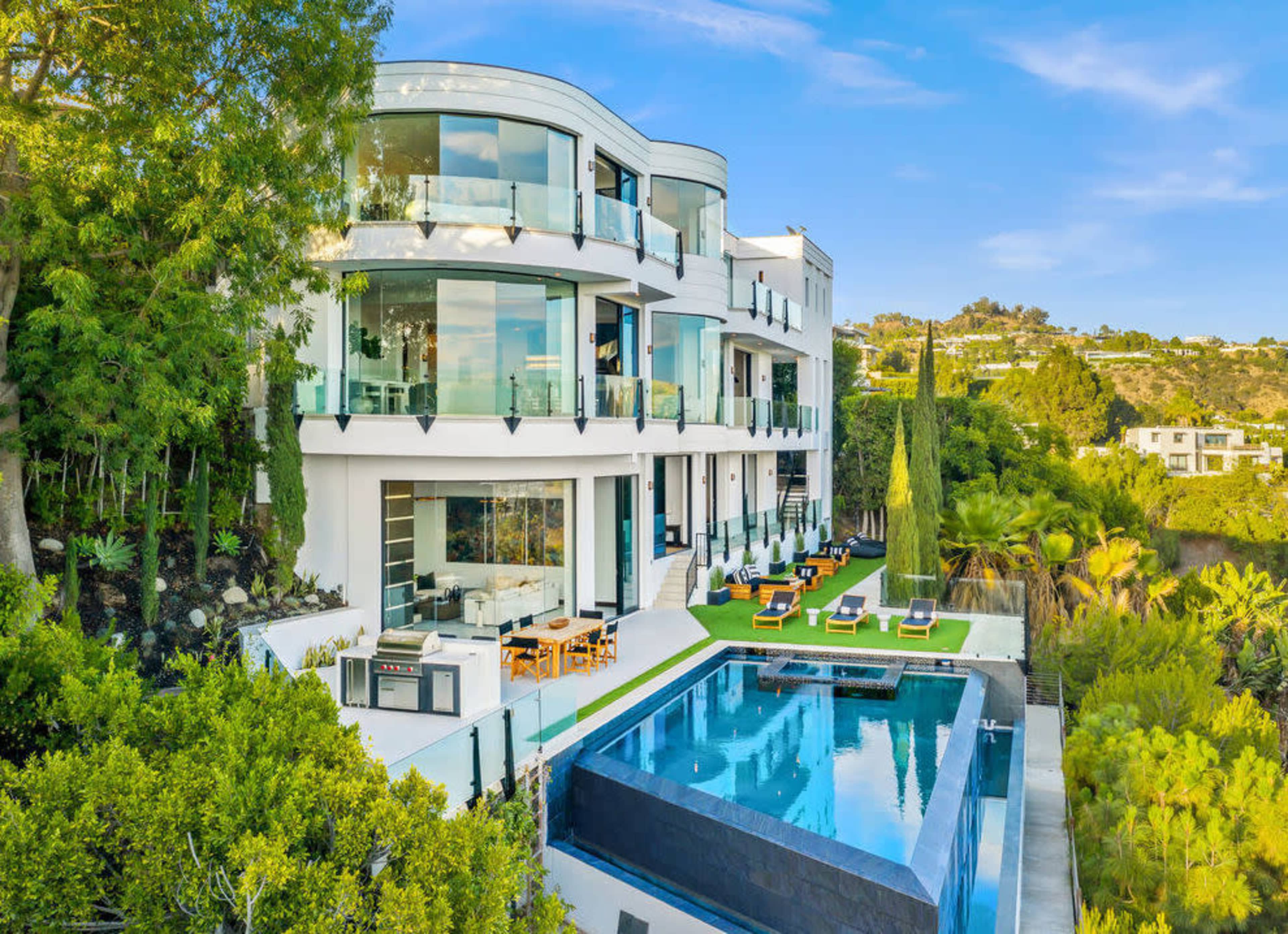 The image shows a modern multi-level house with large glass windows, a swimming pool, and well-manicured gardens.