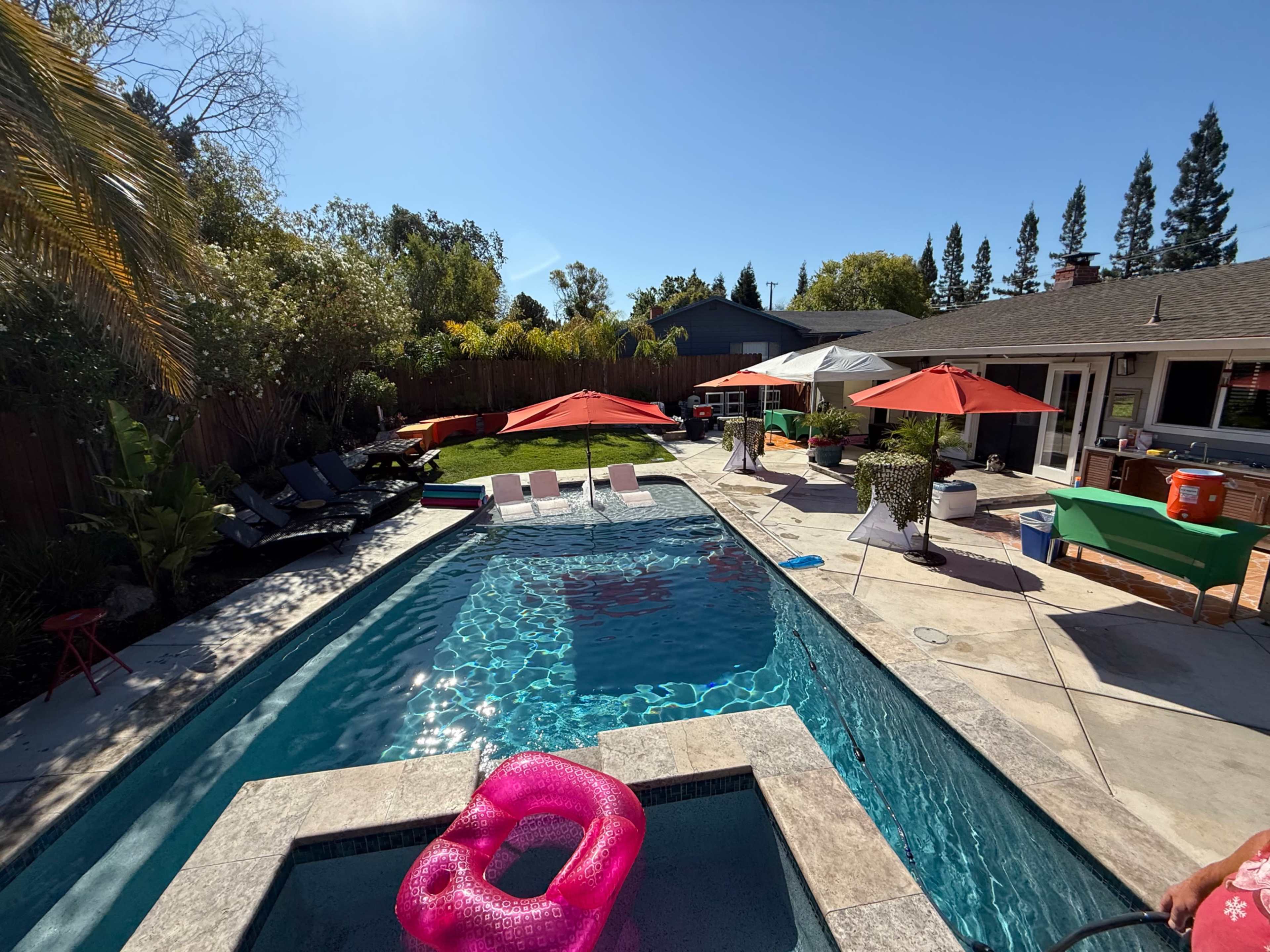 The image shows a backyard pool area with lounge chairs, umbrellas, and a few inflatable floats, surrounded by greenery.