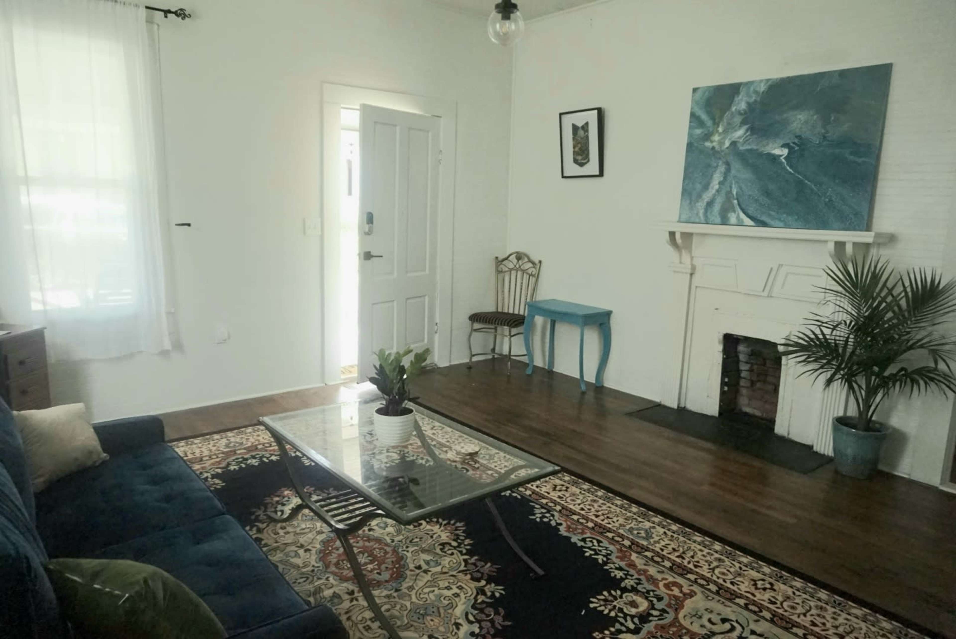 A living room features a blue sofa, a glass coffee table, a plant, and a painting above a white mantel with a fireplace.